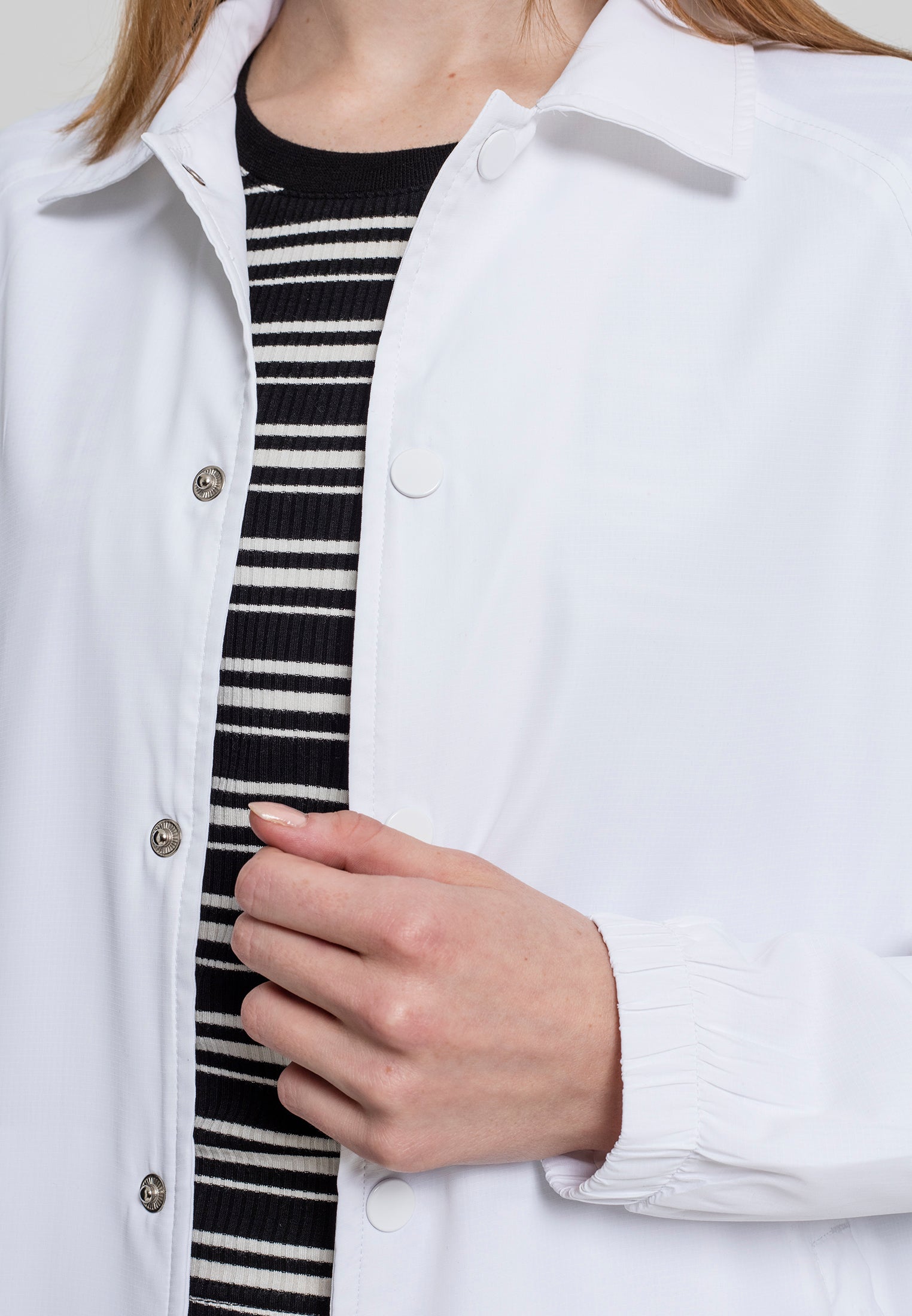 En närbild på en kvinna som bär en vit UC Coach Jacket, med fokus på jackans krage, tryckknappar och elastiska ärmslut.