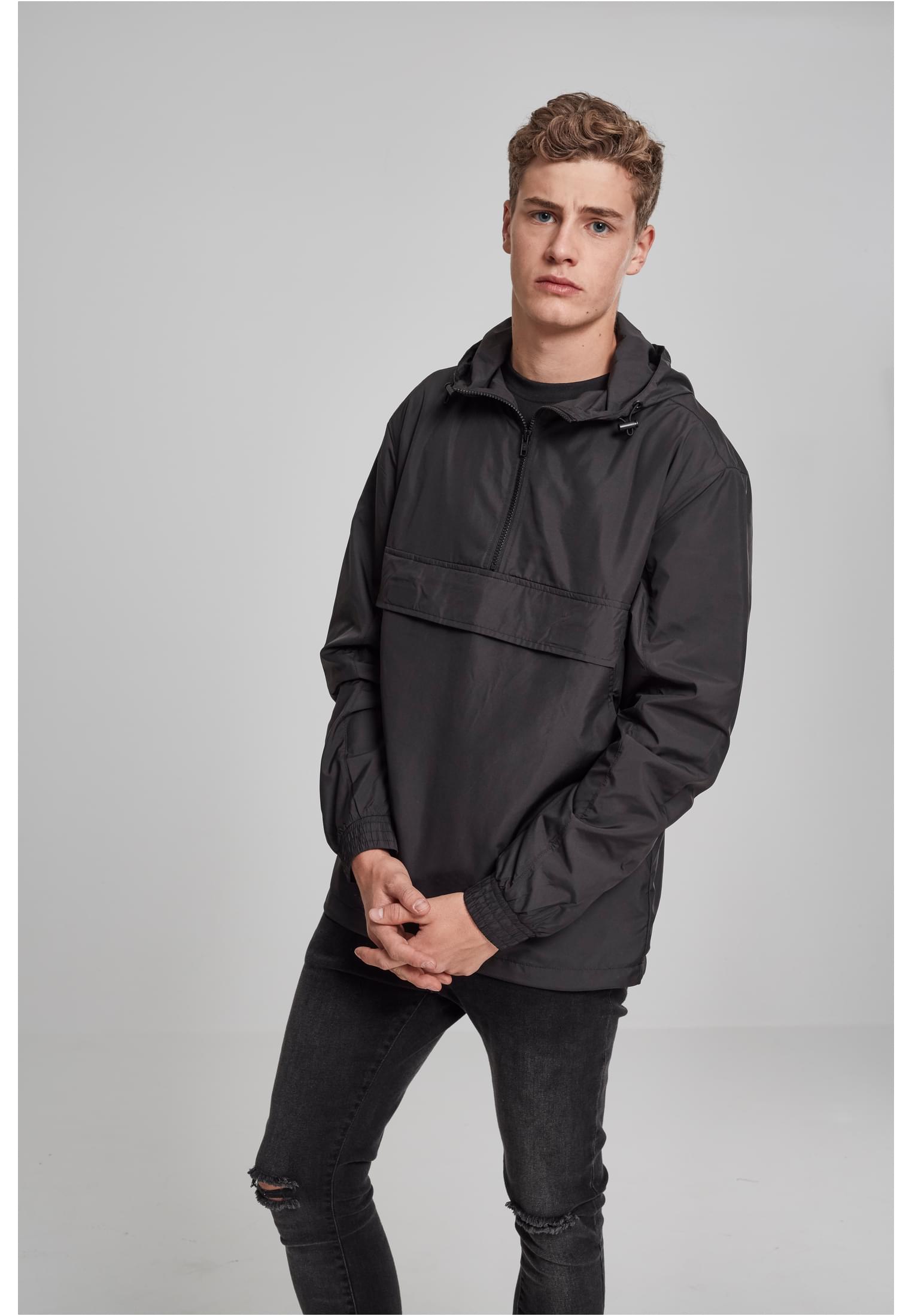 En man poserar i en svart UC Basic Pull Over Jacket och svarta jeans framför en ljusgrå bakgrund.