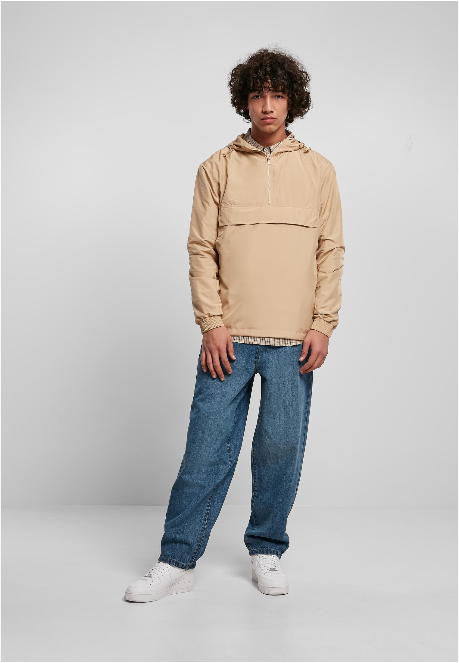 En manlig modell visar framsidan av en beige UC Basic Pull Over Jacket, stylad med blå jeans och vita sneakers, mot en vit bakgrund.