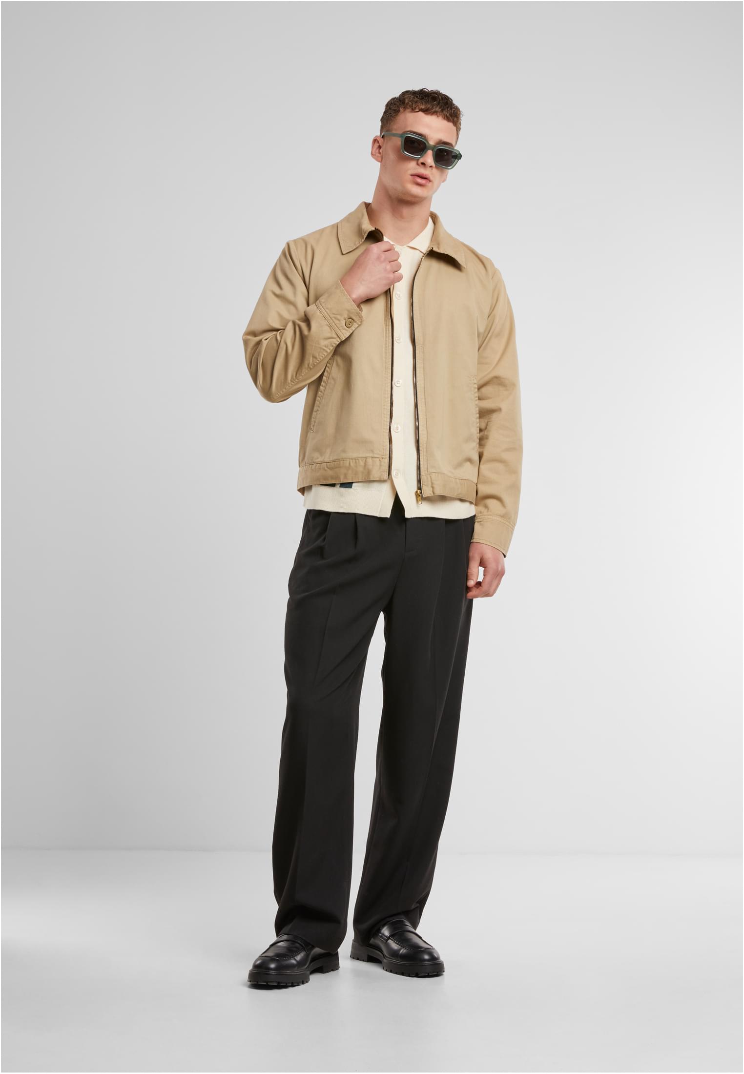 En manlig modell visar framsidan av den beige UC Workwear Jacket, stylad med en ljus skjorta, svarta byxor, svarta skor och solglasögon.