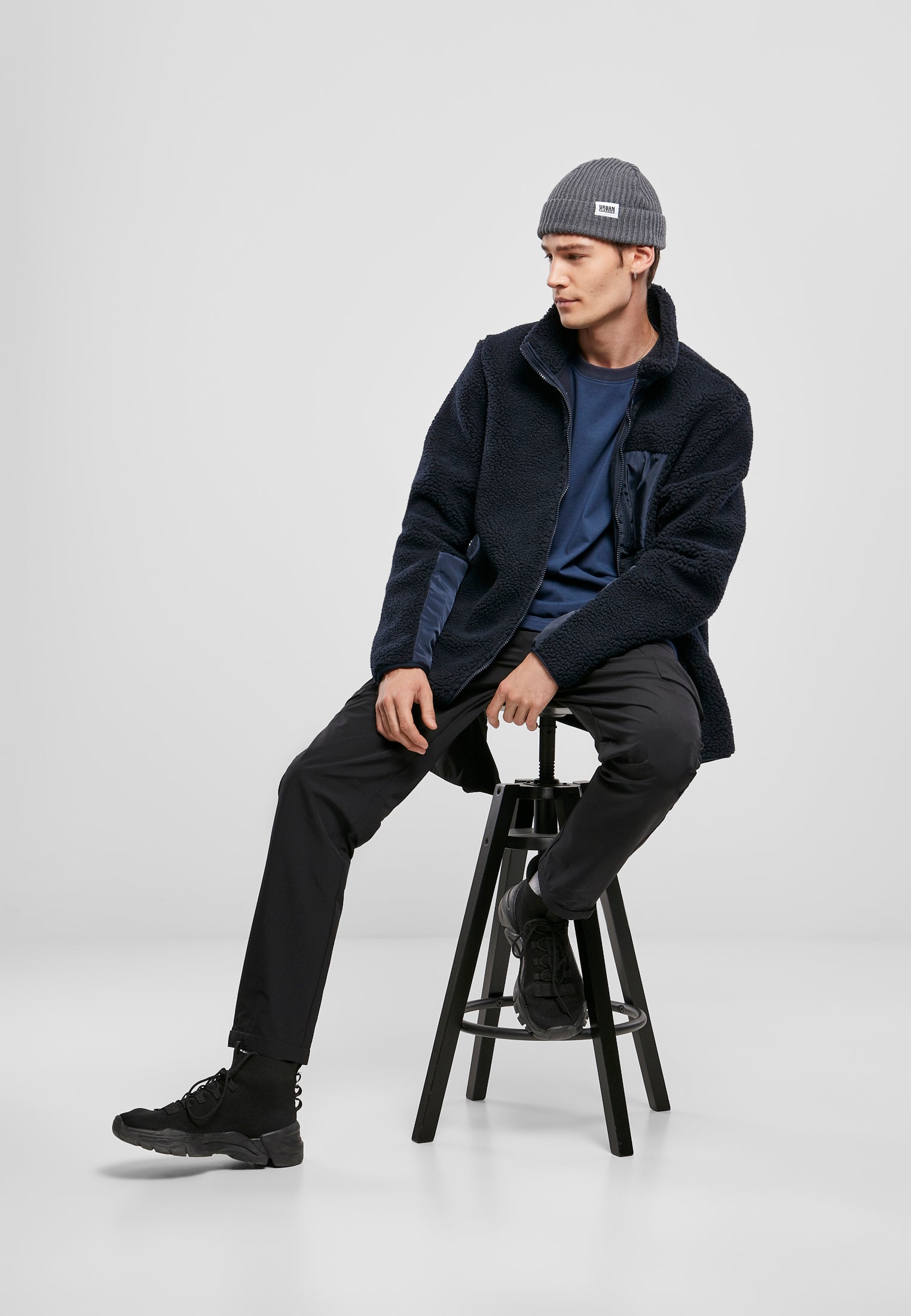 En man som sitter på en pall, iklädd en marinblå UC Sherpa Jacket, en blå t-shirt, svarta byxor och en grå mössa.
