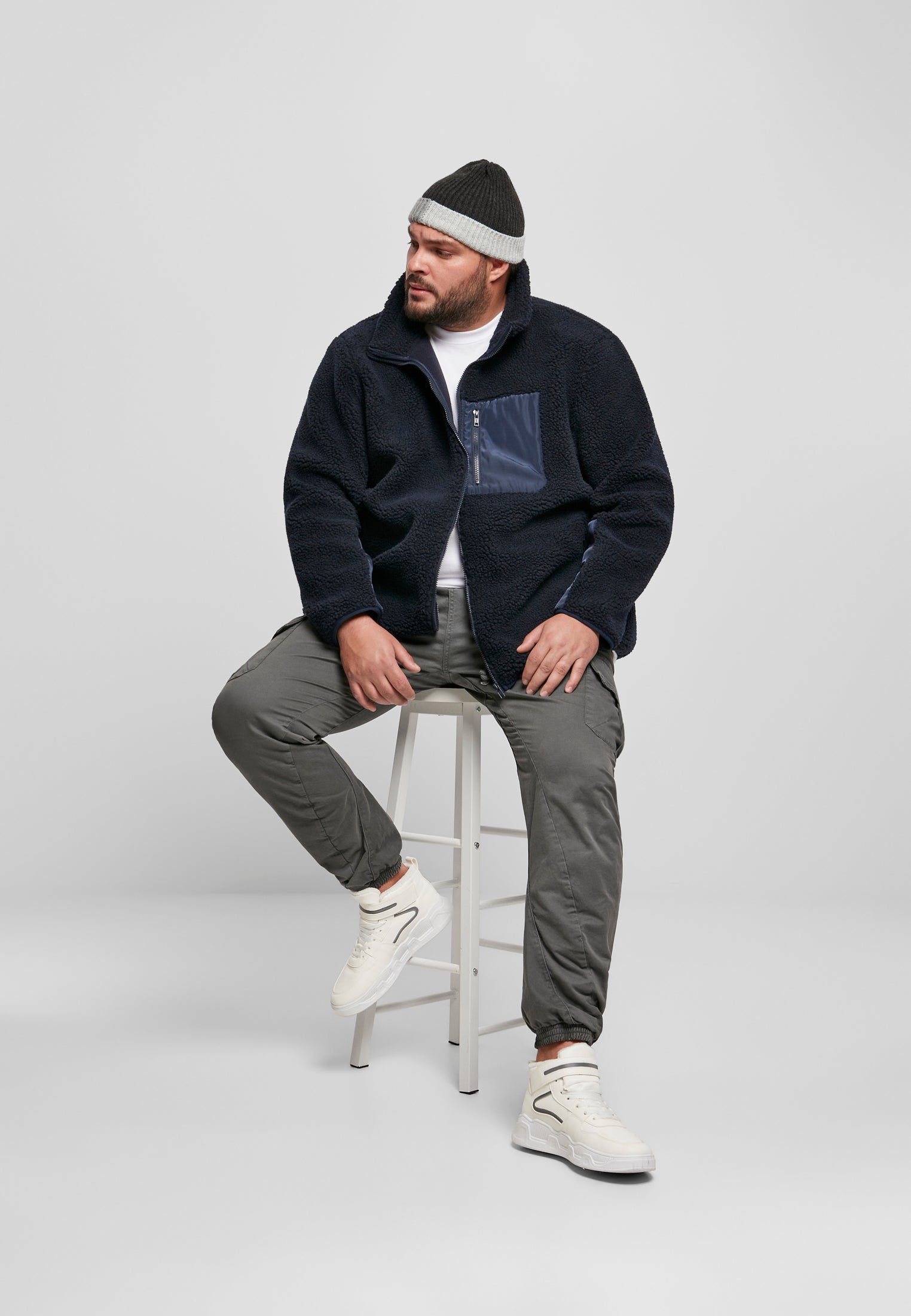 En man sitter på en vit pall och bär en mörkblå UC Sherpa Jacket, gråa byxor, vita sneakers och en mössa.
