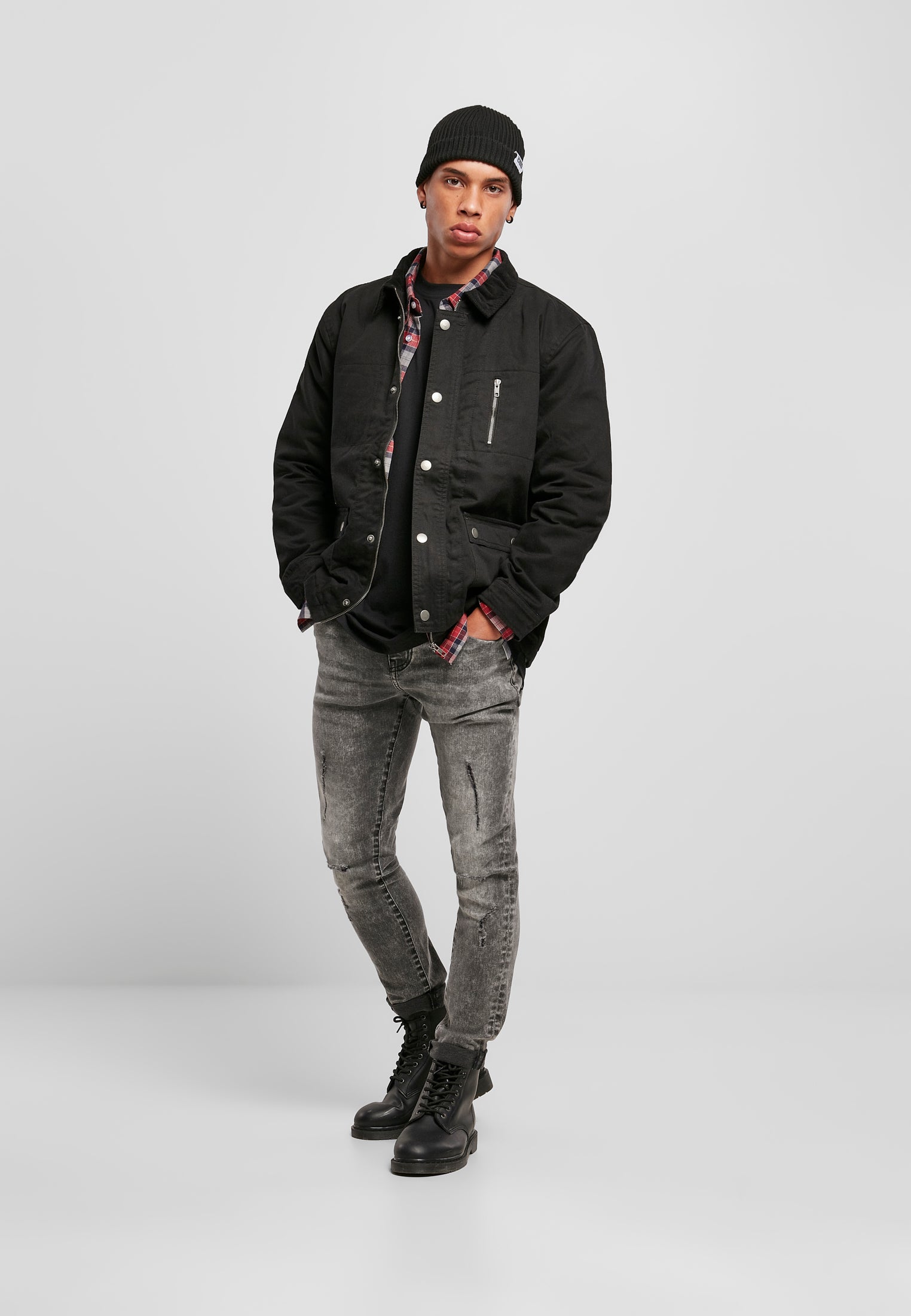 En man poserar och visar framsidan av den svarta jackan UC Hunter Jacket, stylad med en rutig skjorta, mössa, grå jeans och kängor.