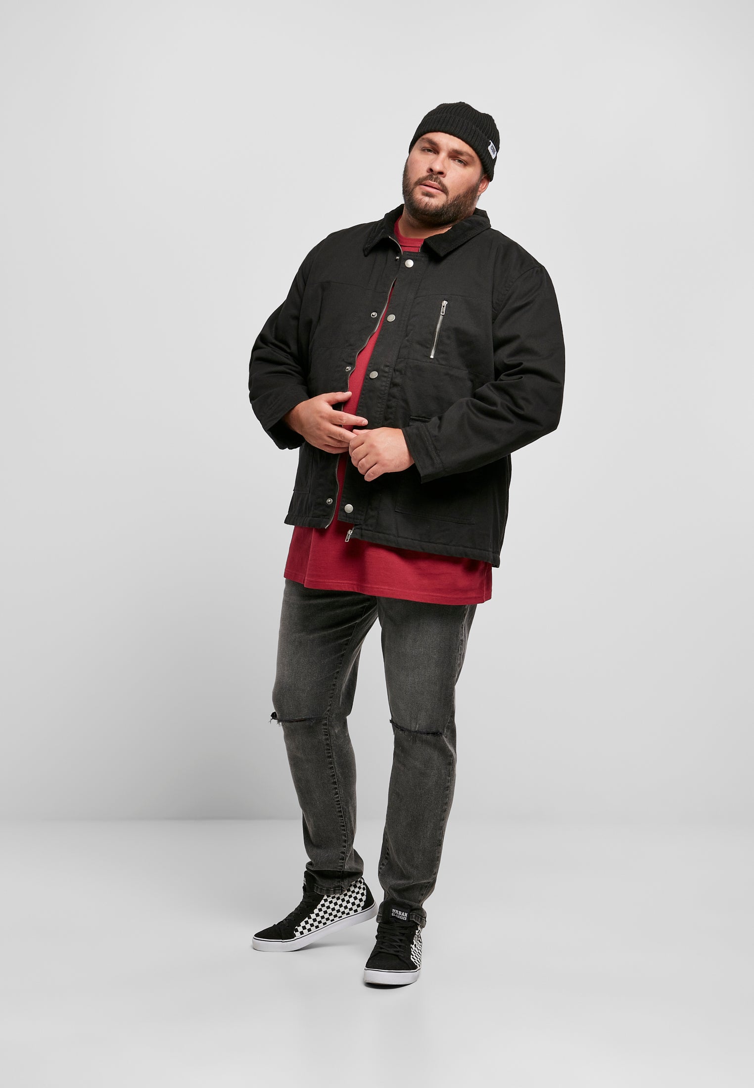 Helkroppsbild på en man som bär en svart UC Hunter Jacket, röd t-shirt, slitna grå jeans och en svart mössa.