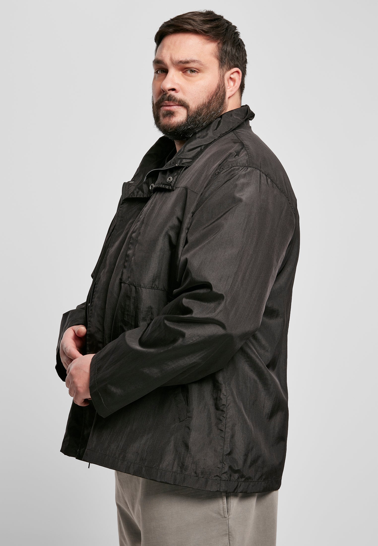 En man med skägg visar sidan av sin svarta UC Double Pocket Nylon Crepe-jacka mot en ljusgrå bakgrund.