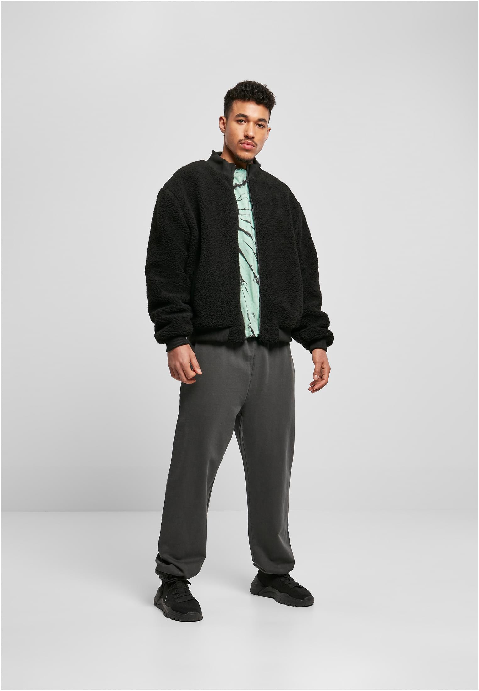 En man visar framsidan av en svart, boxig sherpajacka, buren öppen över en mönstrad t-shirt och mörkgråa byxor.