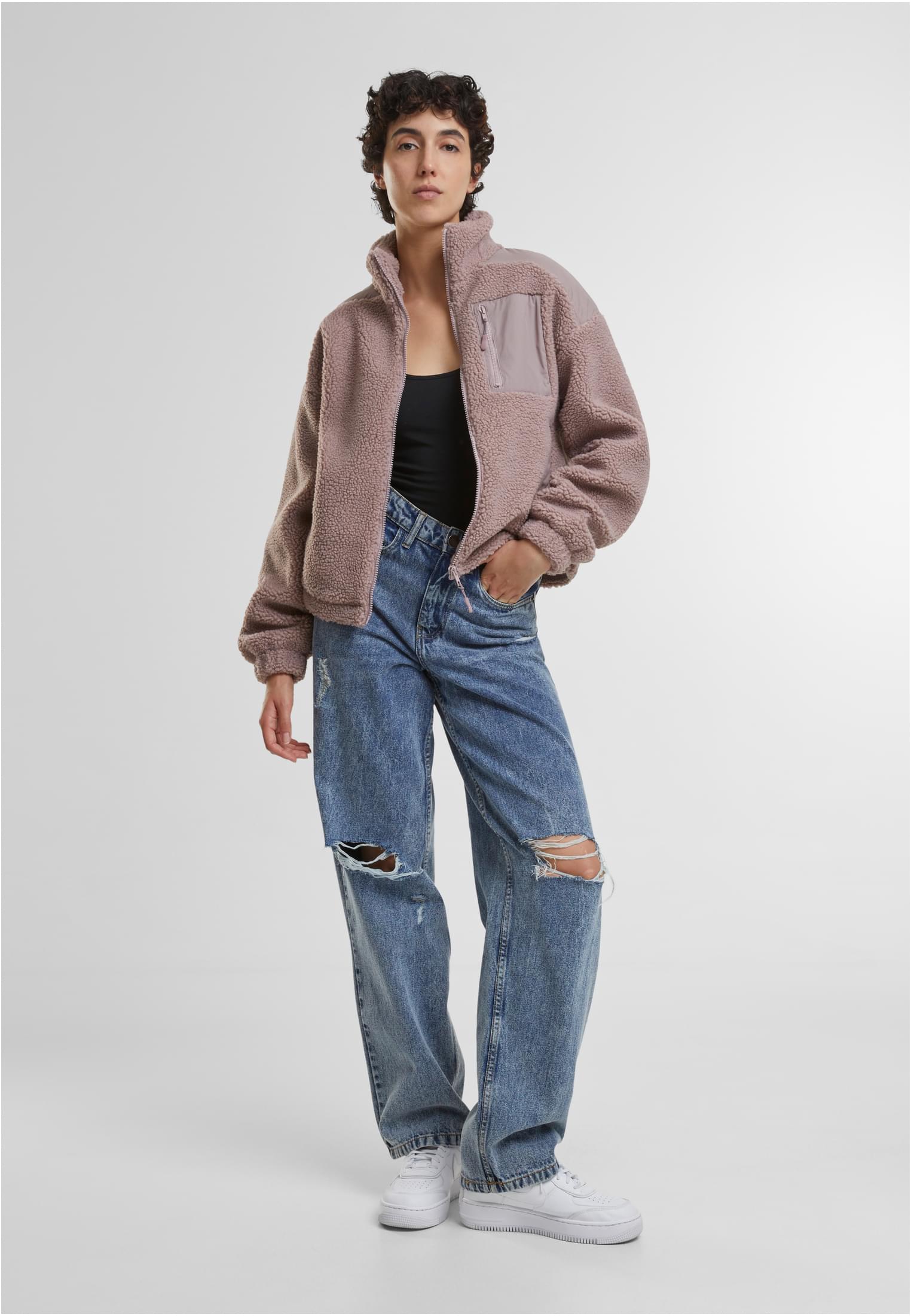 En kvinna som bär en rosa UC Sherpa Mix Jacket, en svart topp och slitna blå jeans.