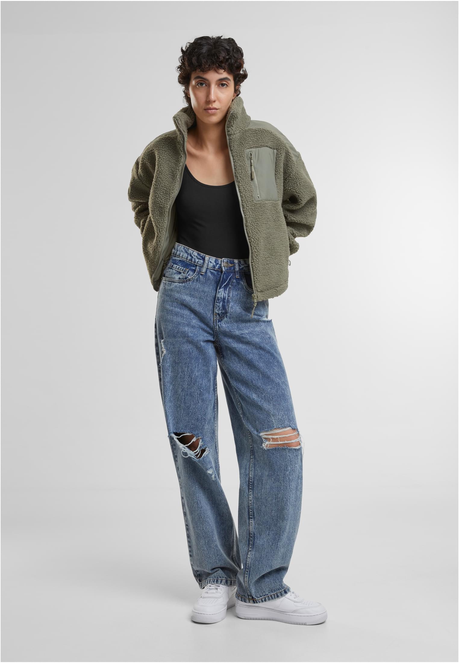 En kvinna poserar i en grön sherpajacka, ett svart linne och slitna blå jeans.