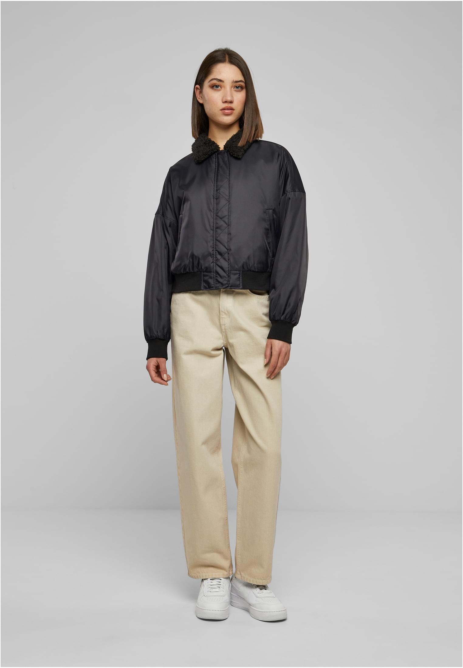 En kvinna visar framsidan av en svart bomberjacka med sherpakrage, stylad med beiga byxor och vita sneakers.