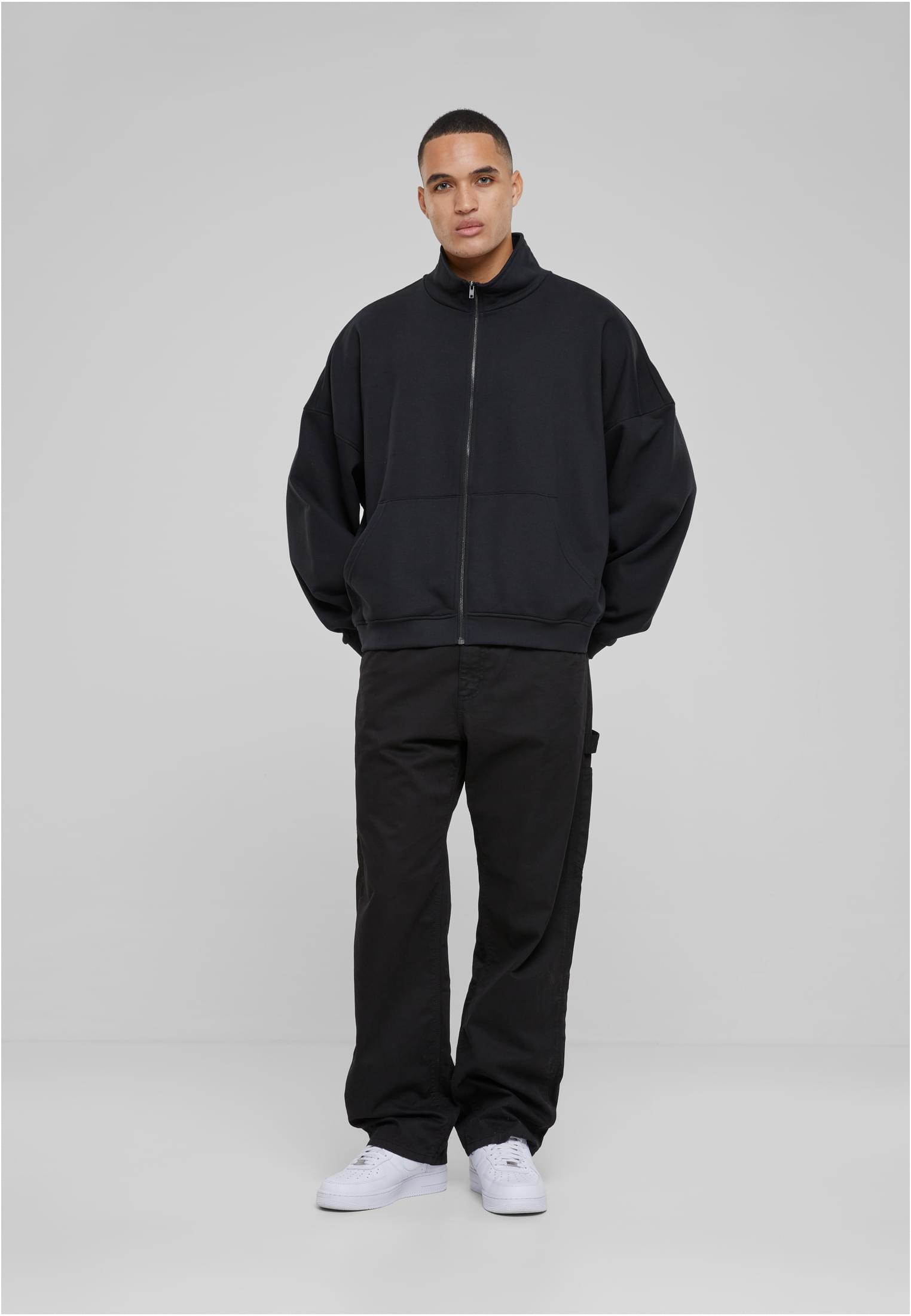 En manlig modell poserar i en svart oversized UC 90s Track Jacket, stylad med svarta byxor och vita sneakers, mot en ljusgrå bakgrund.