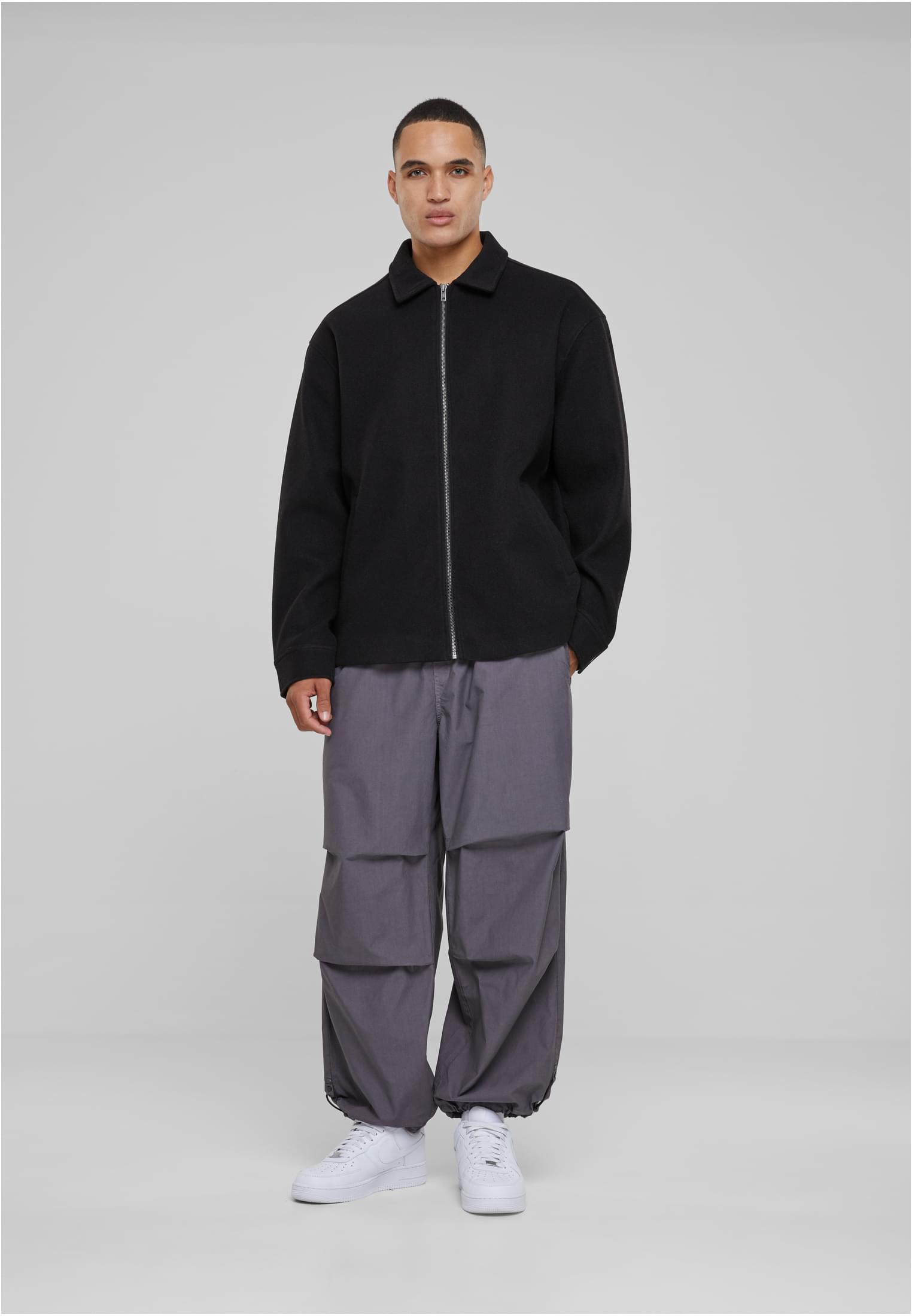 En man visar framsidan av en svart UC Basic Blouson Jacket, buren med gråa byxor och vita sneakers.