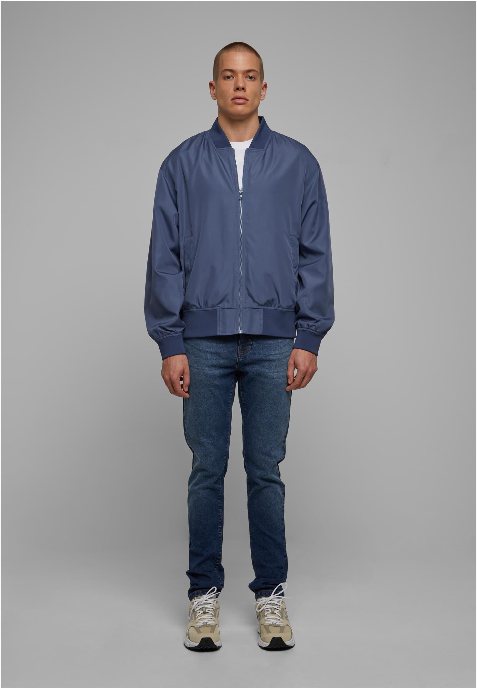 En man, sedd framifrån, bär en blå bomberjacka, vit t-shirt och jeans mot en grå bakgrund.