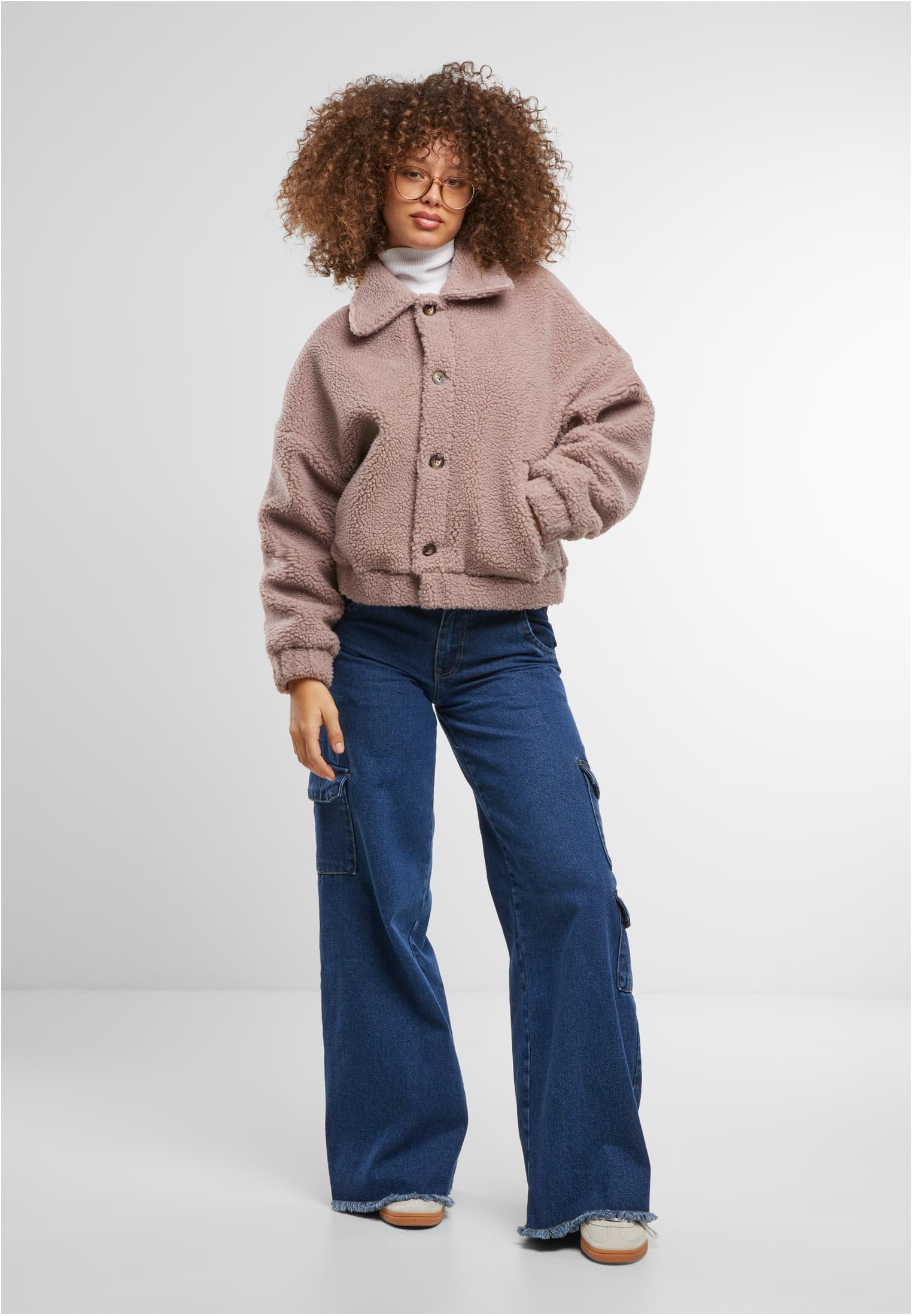 En kvinna med lockigt hår och glasögon bär en rosa UC Short Sherpa Button Jacket, en vit polotröja och vida jeans.