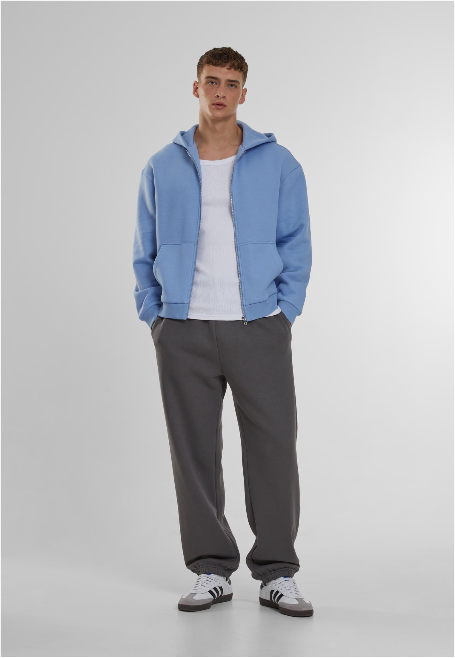 En manlig modell poserar i en helkroppsbild iklädd mörkgråa mjukisbyxor, en ljusblå huvtröja med dragkedja och ett vitt linne.