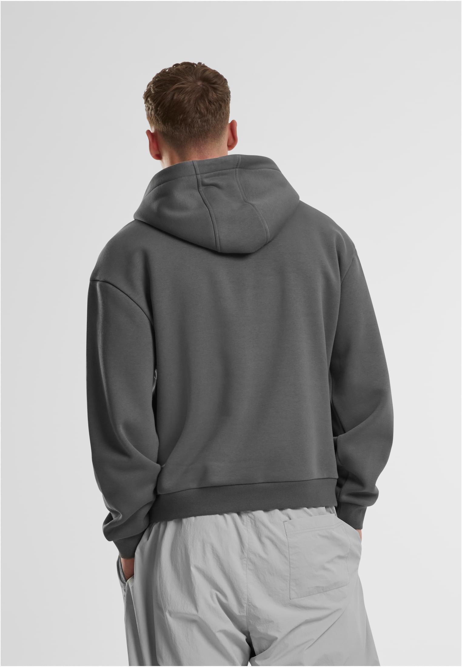 En man sedd bakifrån som bär en mörkgrå luvtröja och ljusgrå byxor, stående mot en vit bakgrund.