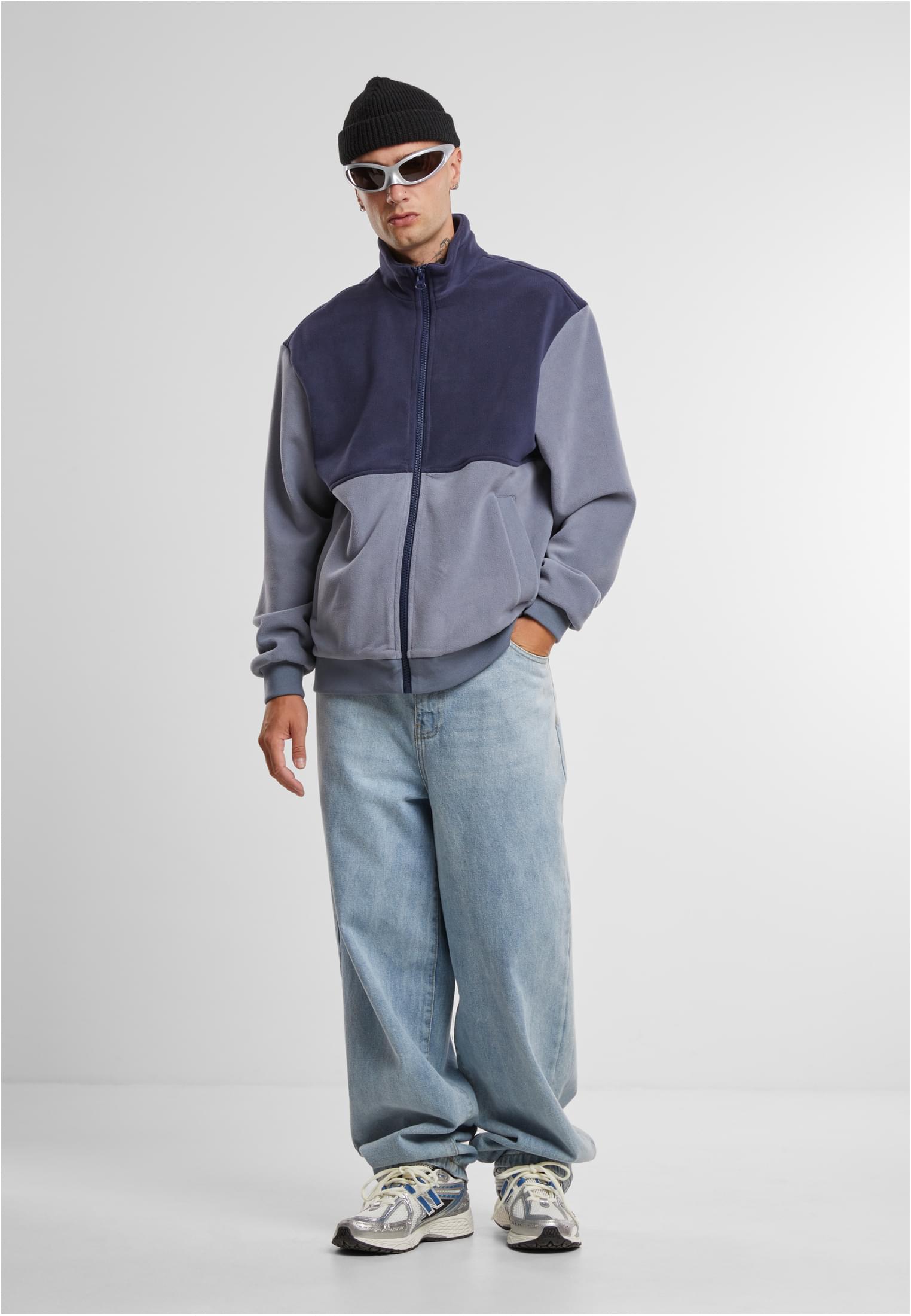 En man poserar i en tvåfärgad blå fleecejacka med dragkedja, kombinerad med vida jeans, en svart mössa och silverfärgade solglasögon.