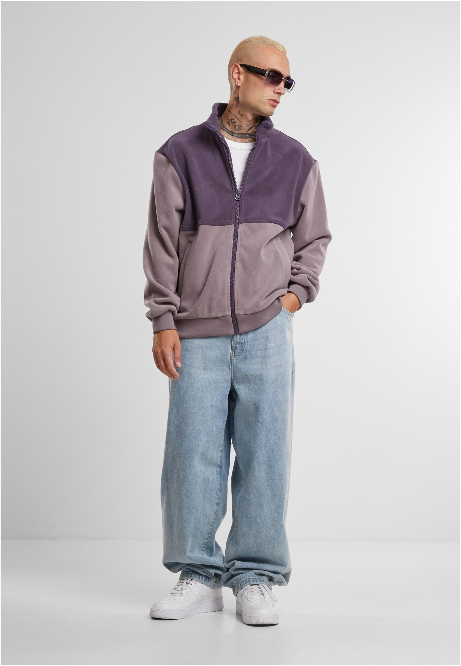 En man med solglasögon och tatueringar bär en tvåfärgad lila fleecejacka med dragkedja, stylad med vida jeans och vita sneakers.