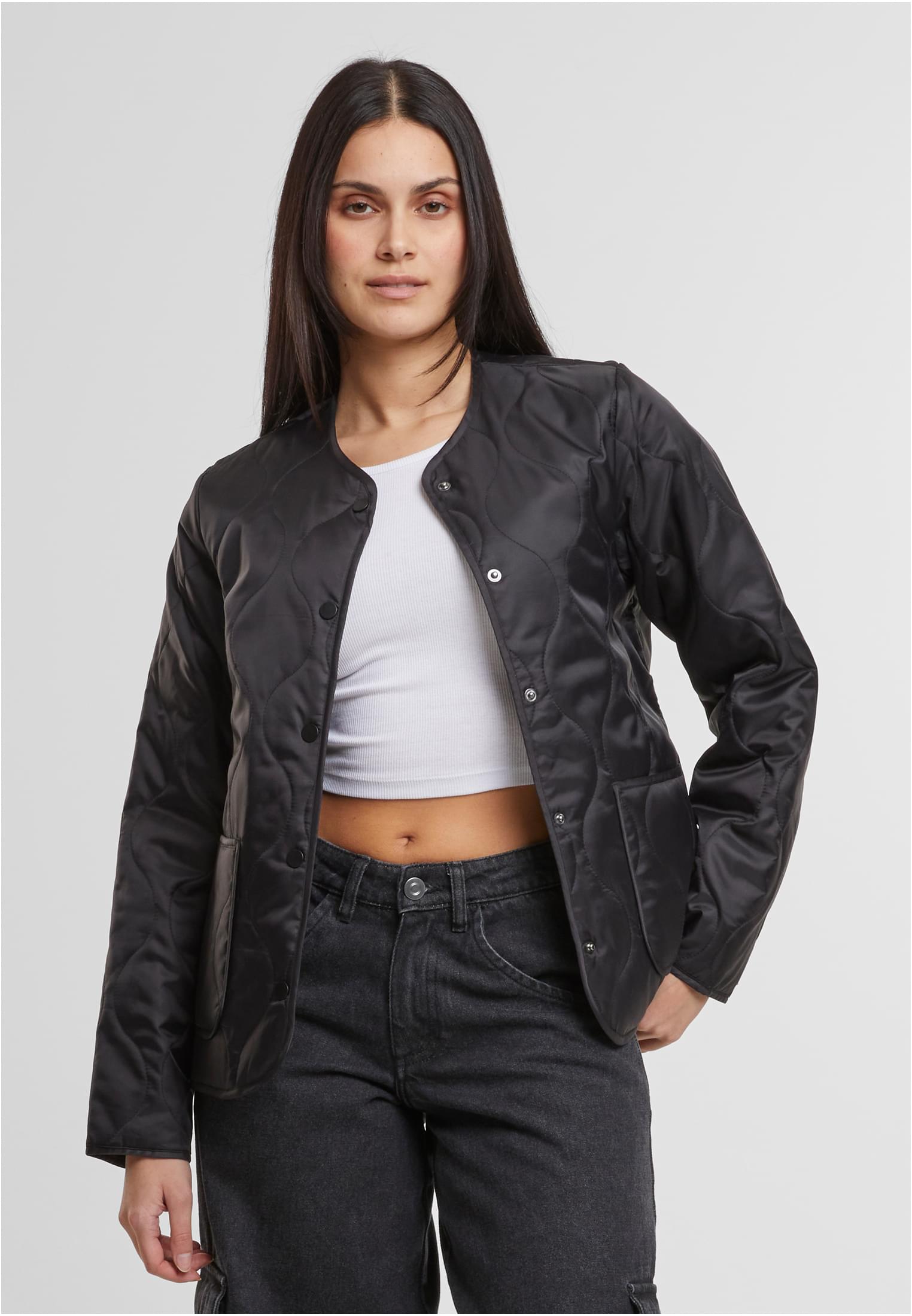 En kvinna som bär en svart, quiltad jacka över en vit topp och mörkgråa jeans, fotograferad framifrån.