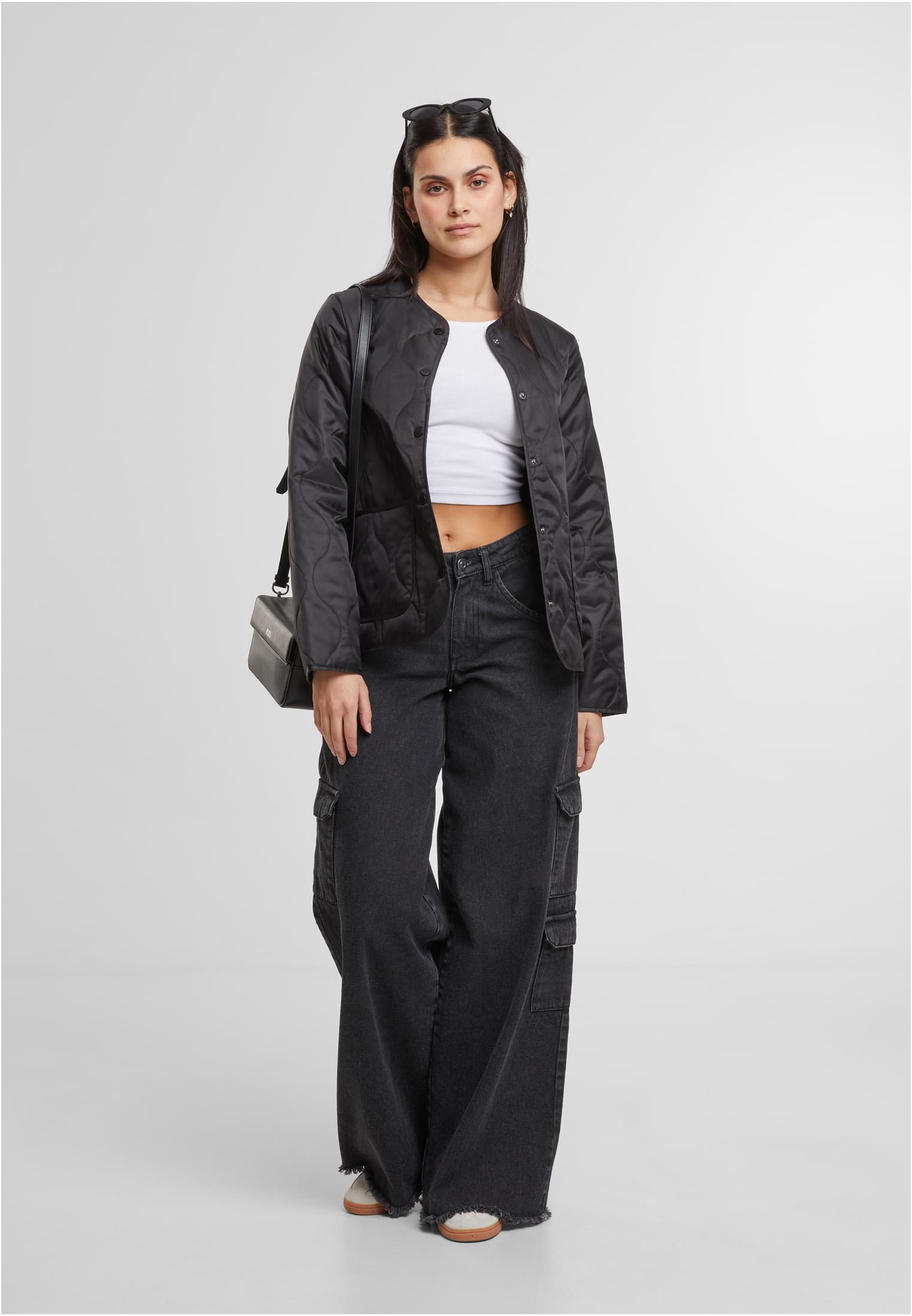 En kvinna poserar framifrån klädd i en svart quiltad jacka, en vit crop top och vida svarta cargojeans.