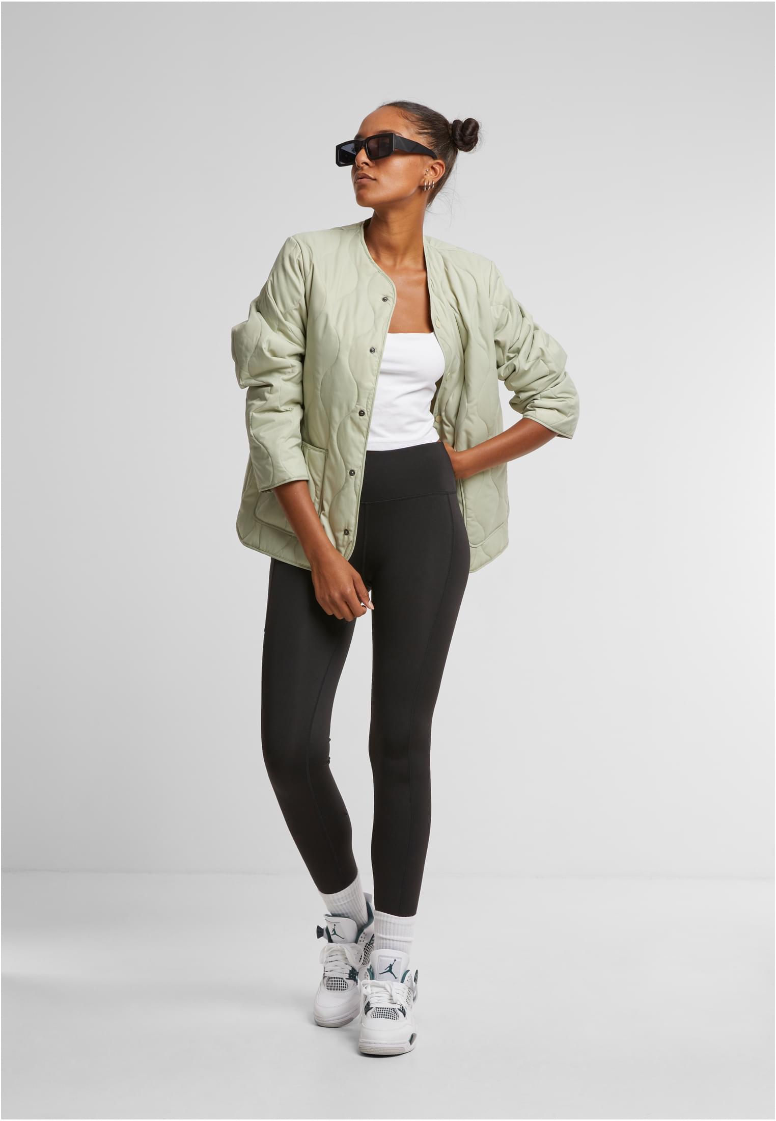 En kvinna bär en ljusgrön quiltad jacka, ett vitt linne, svarta leggings och vita sneakers.
