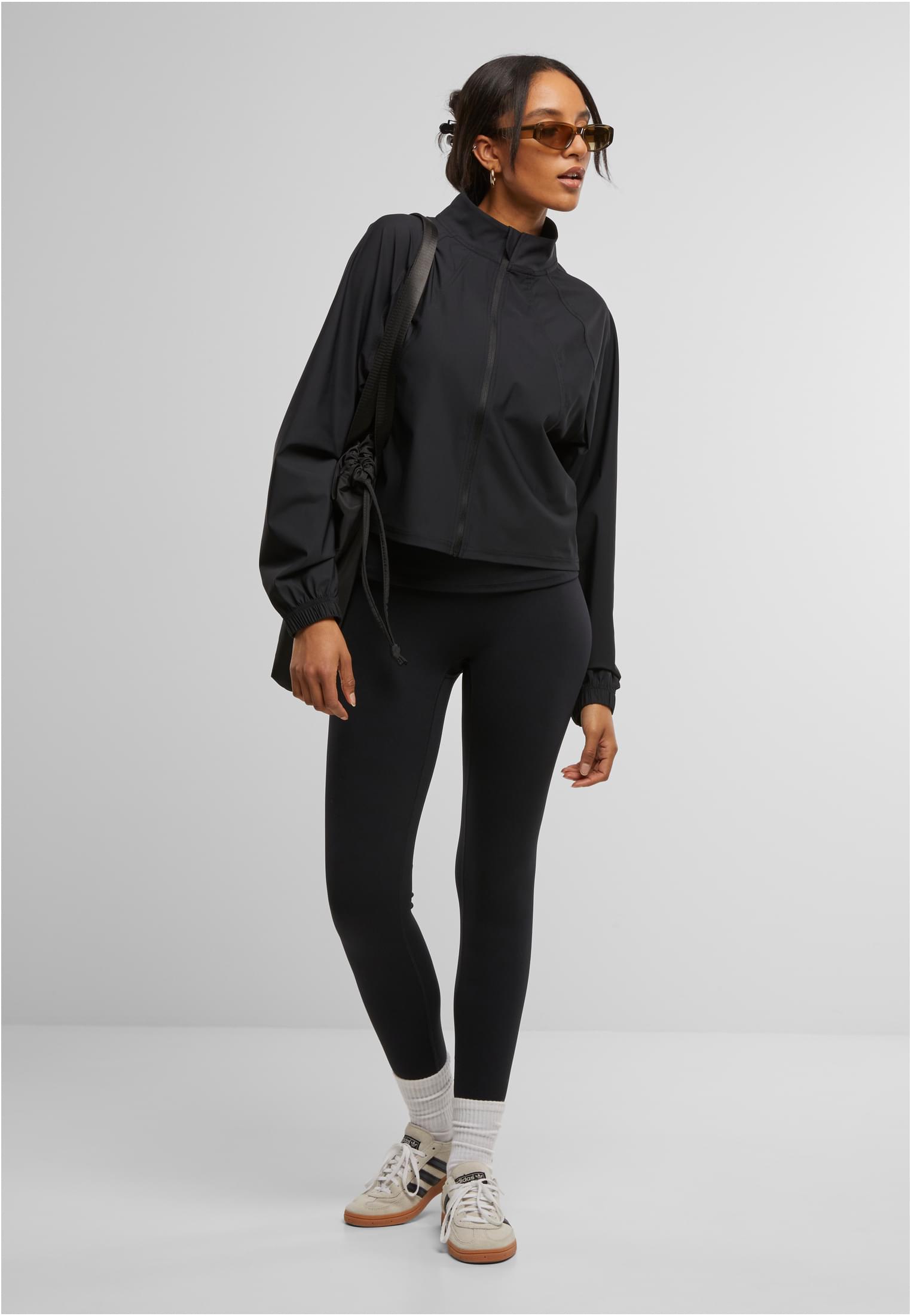 En kvinna i helfigur som bär en svart sportjacka, svarta leggings, ljusa sneakers och solglasögon.