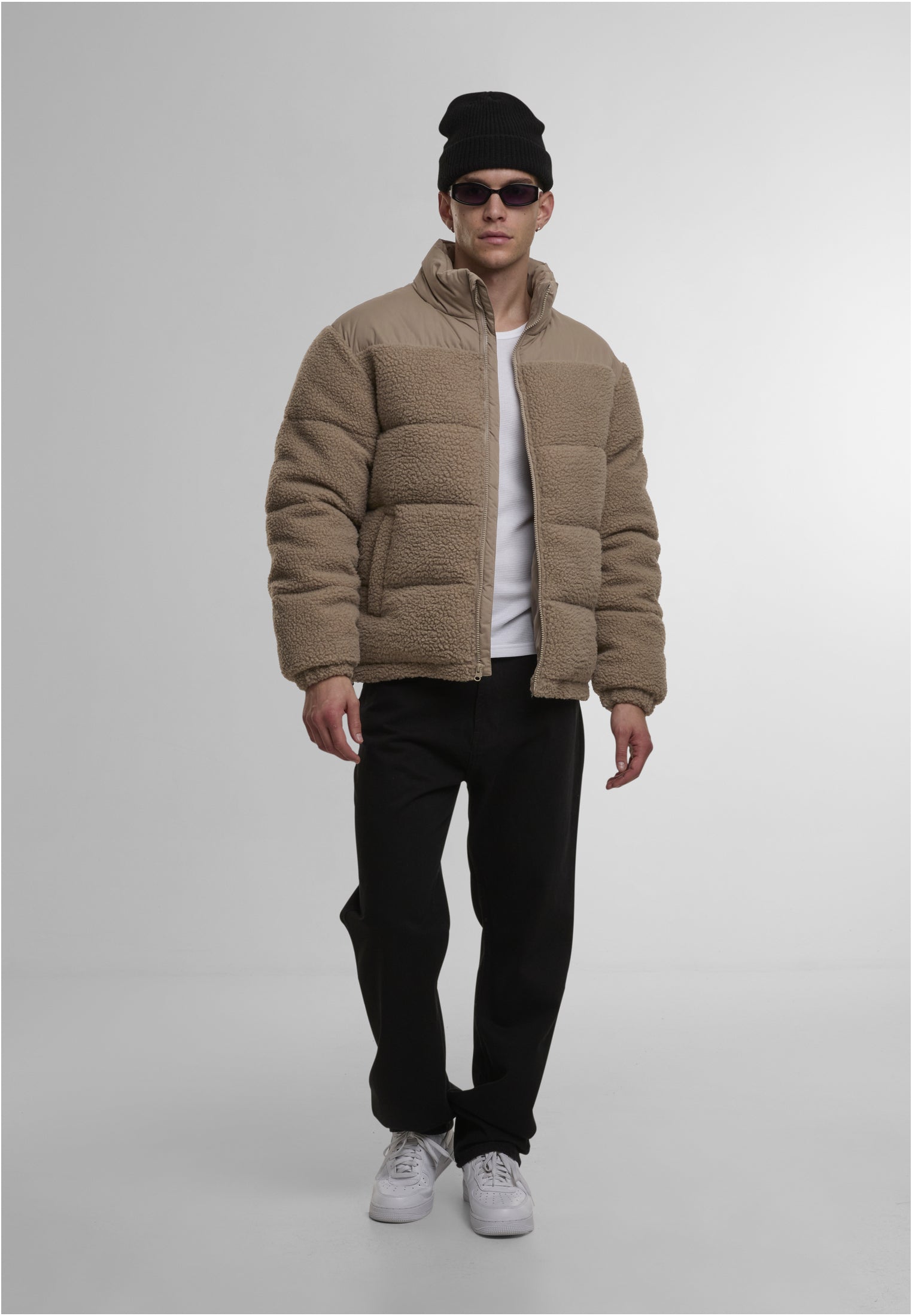 En man fotograferad framifrån i en beige teddy-puffjacka, vit t-shirt, svarta byxor och svart mössa.