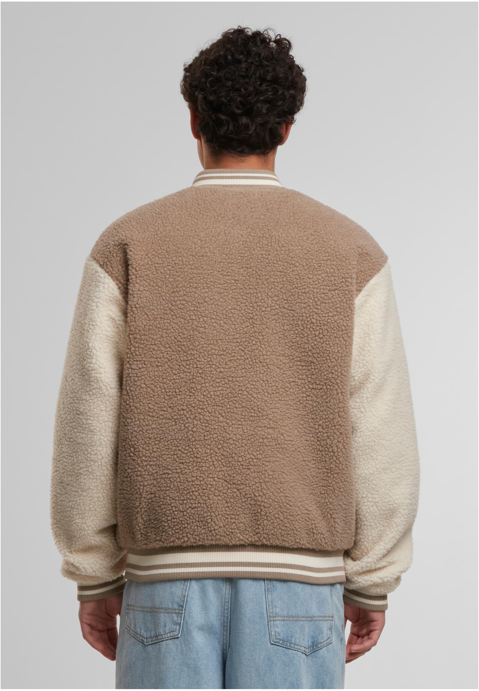 Baksidan av en man som bär en mörkbeige teddy-collegejacka med benvita ärmar och blå jeans.