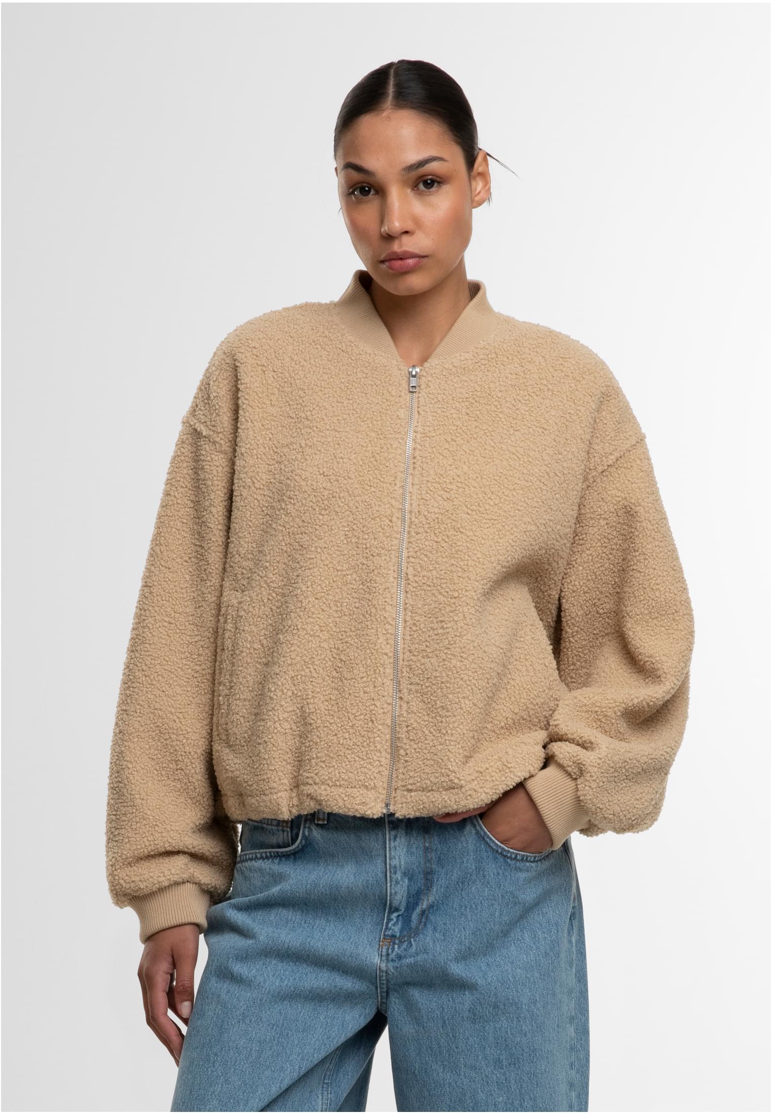 En kvinna som bär en beige bomberjacka i teddymaterial och blå jeans, fotograferad framifrån mot en vit bakgrund.