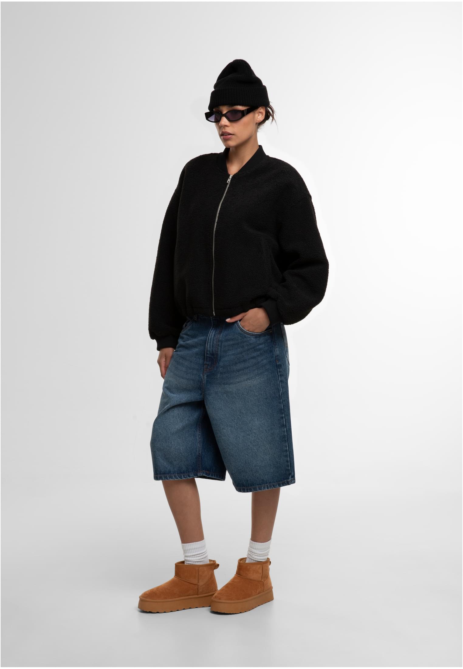 En kvinna i helfigur bär en svart strukturerad bomberjacka, långa jeansshorts, en svart mössa och solglasögon.