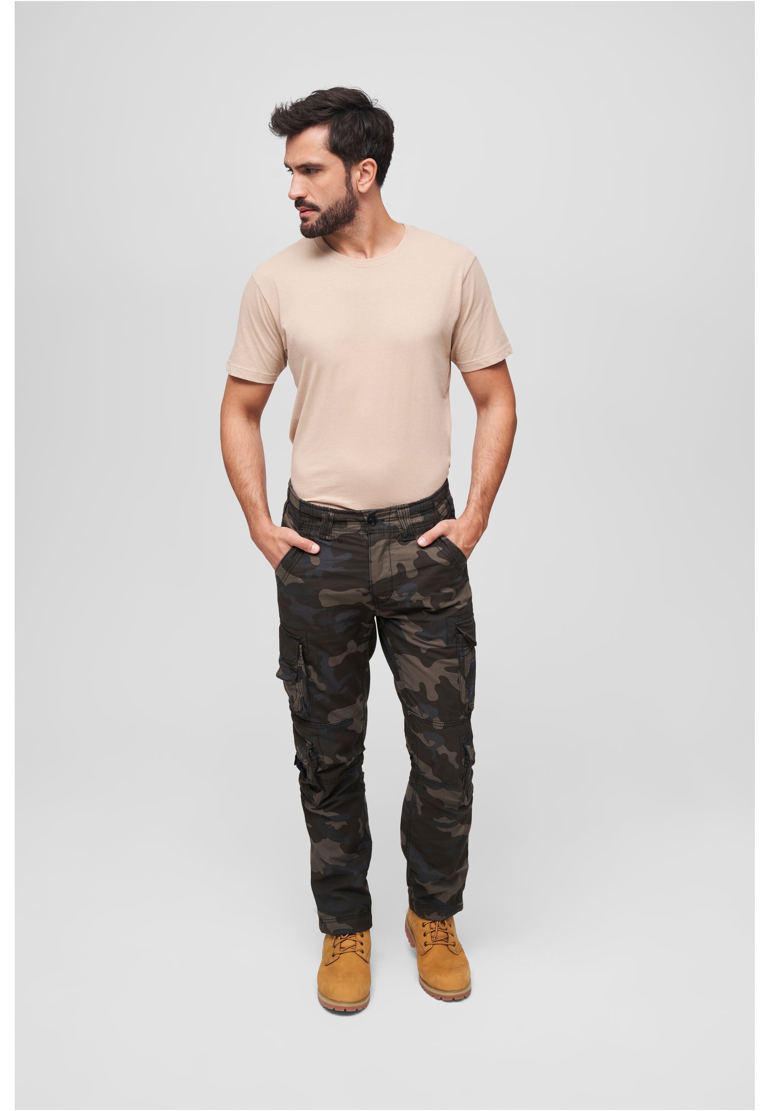 En man med skägg bär en beige t-shirt, kamouflagecargobyxor och ljusbruna kängor, fotograferad i helfigur mot en ljusgrå bakgrund.