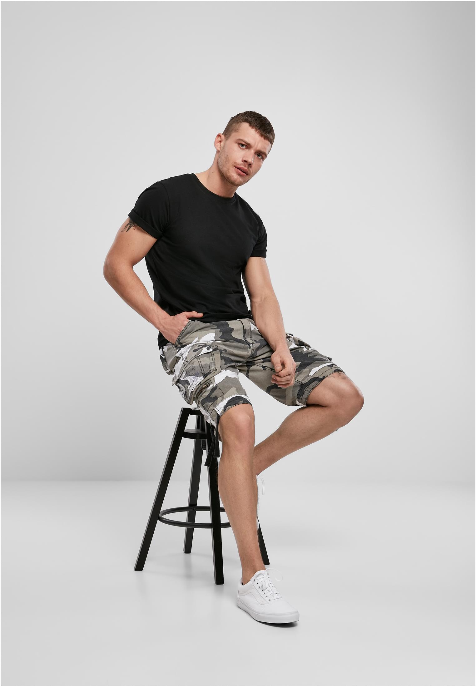 En man sitter på en svart pall och bär en svart B Premium Shirt, kamouflageshorts och vita sneakers.