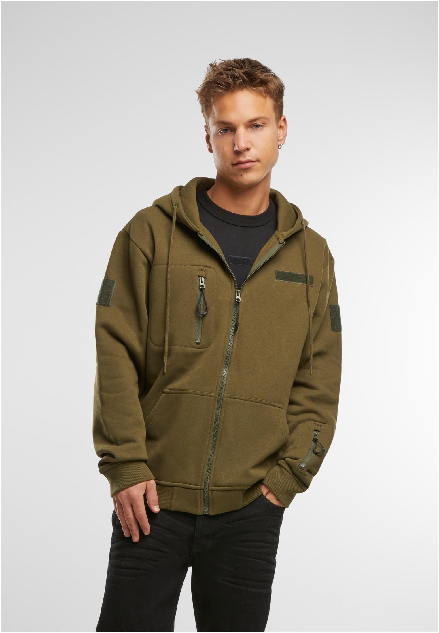 En man visar framsidan av den olivgröna B Tactical Sweat Jacket. Han bär den öppen över en svart t-shirt och svarta jeans.