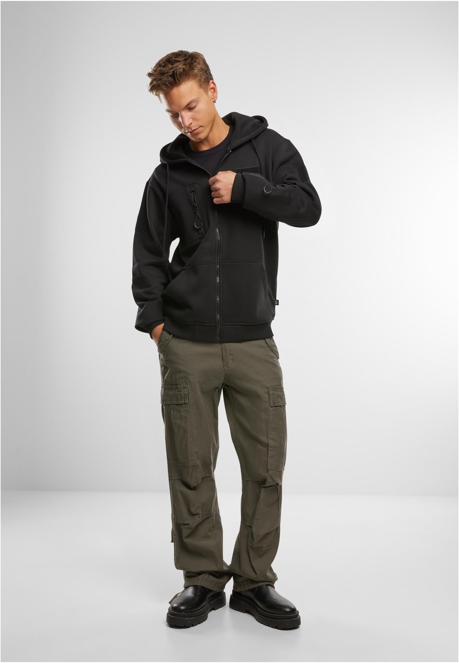 En man fotograferad framifrån, klädd i en svart B Tactical Sweat Jacket, olivgröna cargobyxor och svarta kängor.