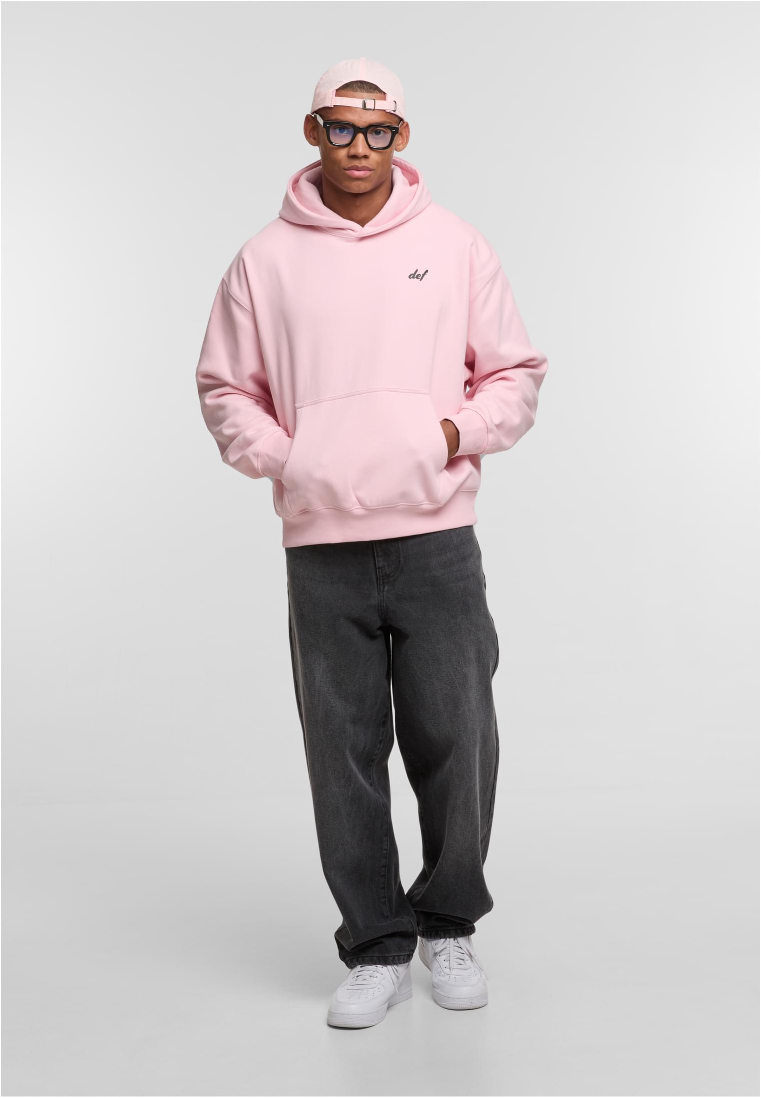 En manlig modell poserar i en rosa DEF VISIONS Hoodie, mörkgråa baggy jeans och en matchande rosa keps.
