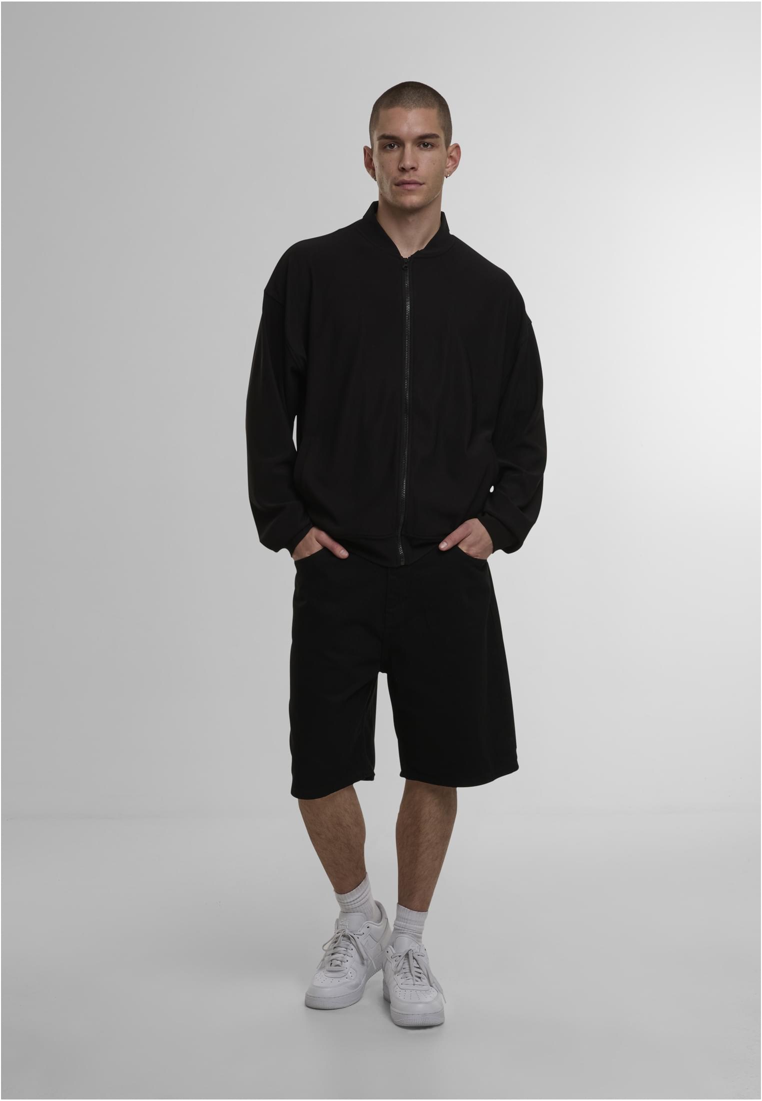 En man visar upp framsidan av den svarta DEF Milan-jackan, stylad med svarta shorts och vita sneakers.