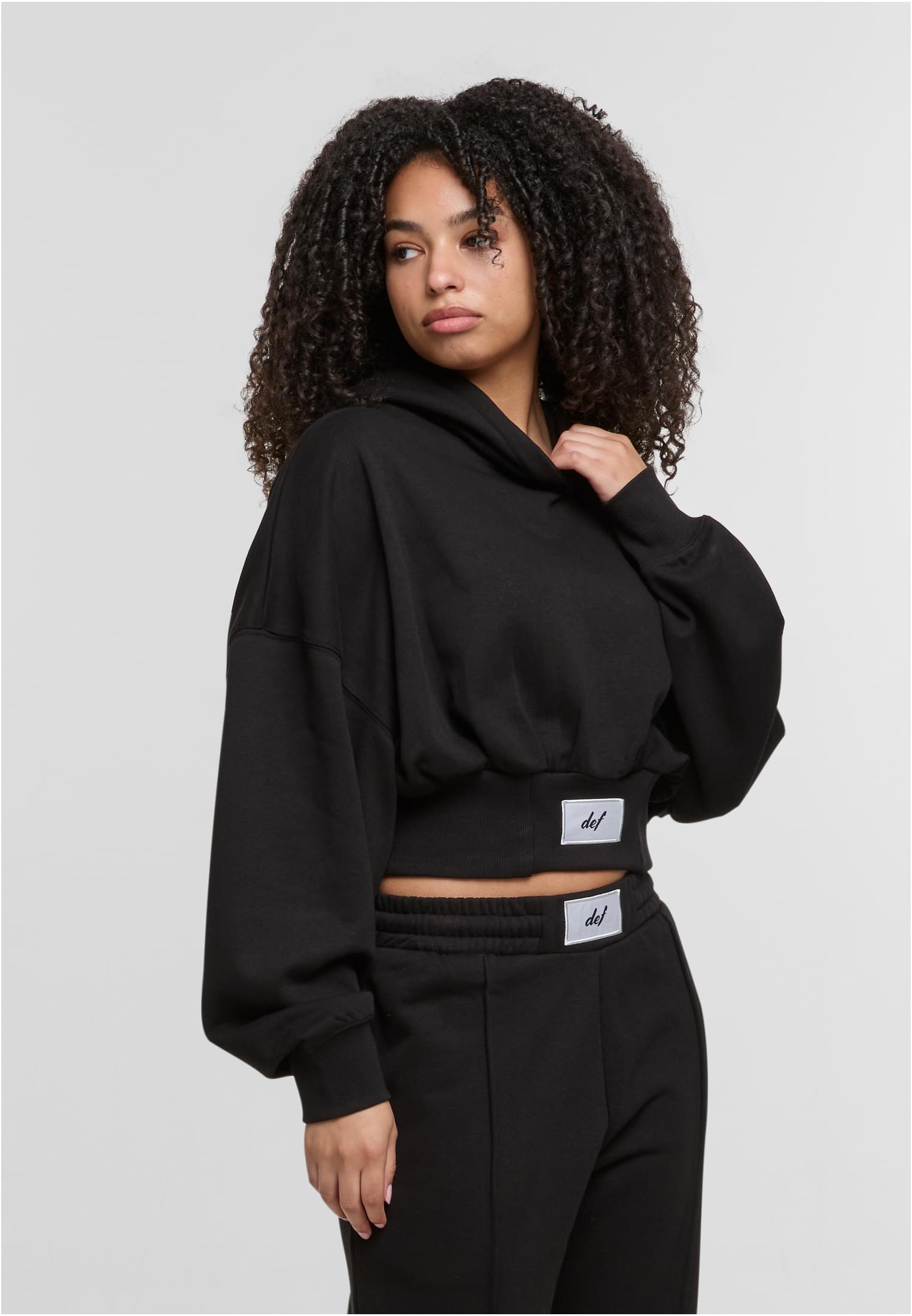 En kvinna som tittar över axeln, klädd i en svart, kort luvtröja och matchande mjukisbyxor från DEF.