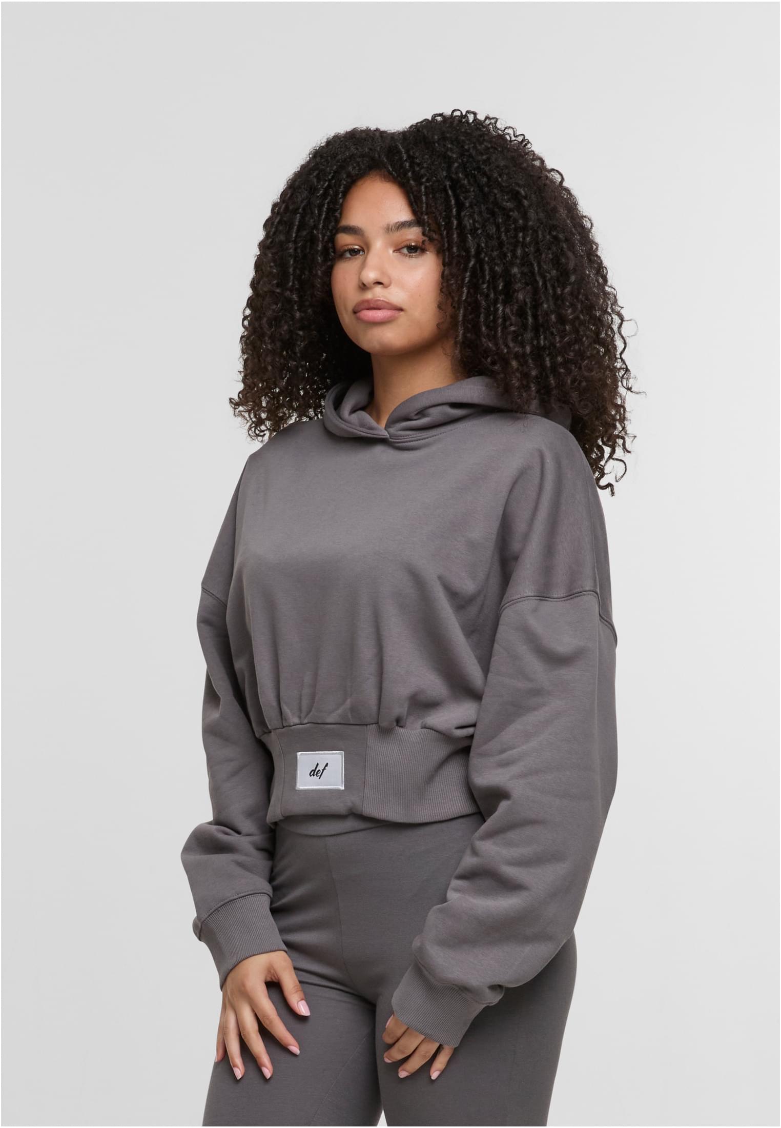 En kvinna med lockigt hår visar framsidan av en grå, croppad DEF NICE Hoodie.