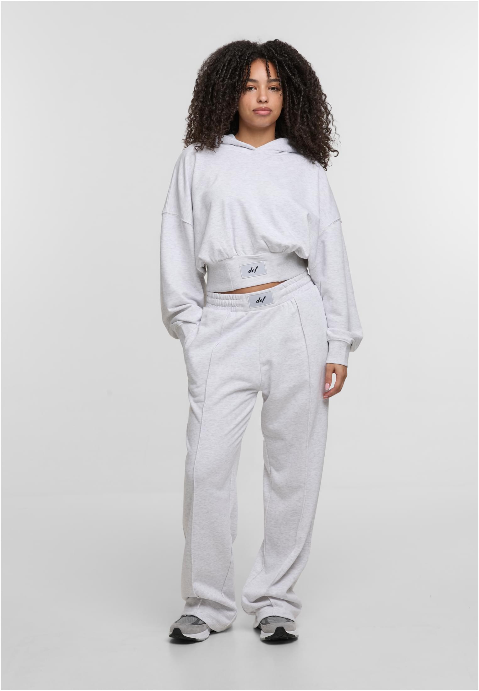 En kvinna som poserar i en ljusgrå, kort DEF NICE Hoodie och matchande mjukisbyxor med vida ben mot en vit bakgrund.