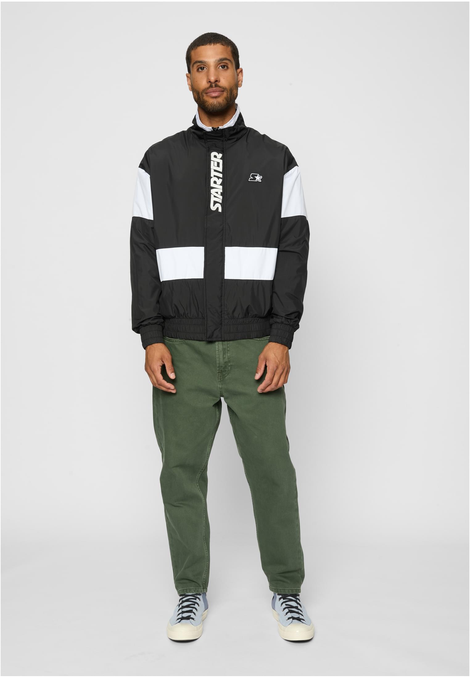 En man visar framsidan av SBL Retro Track Jacket i svart och vitt, stylad med gröna byxor och ljusblå sneakers.