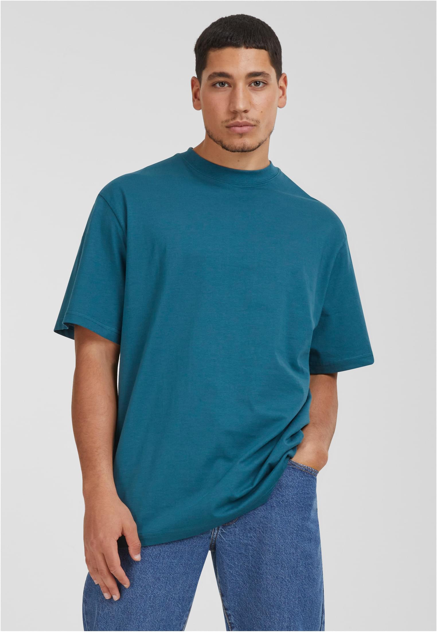 Manlig modell som bär en blågrön UC Tall Tee och blå jeans, fotograferad framifrån mot en ljusgrå bakgrund.