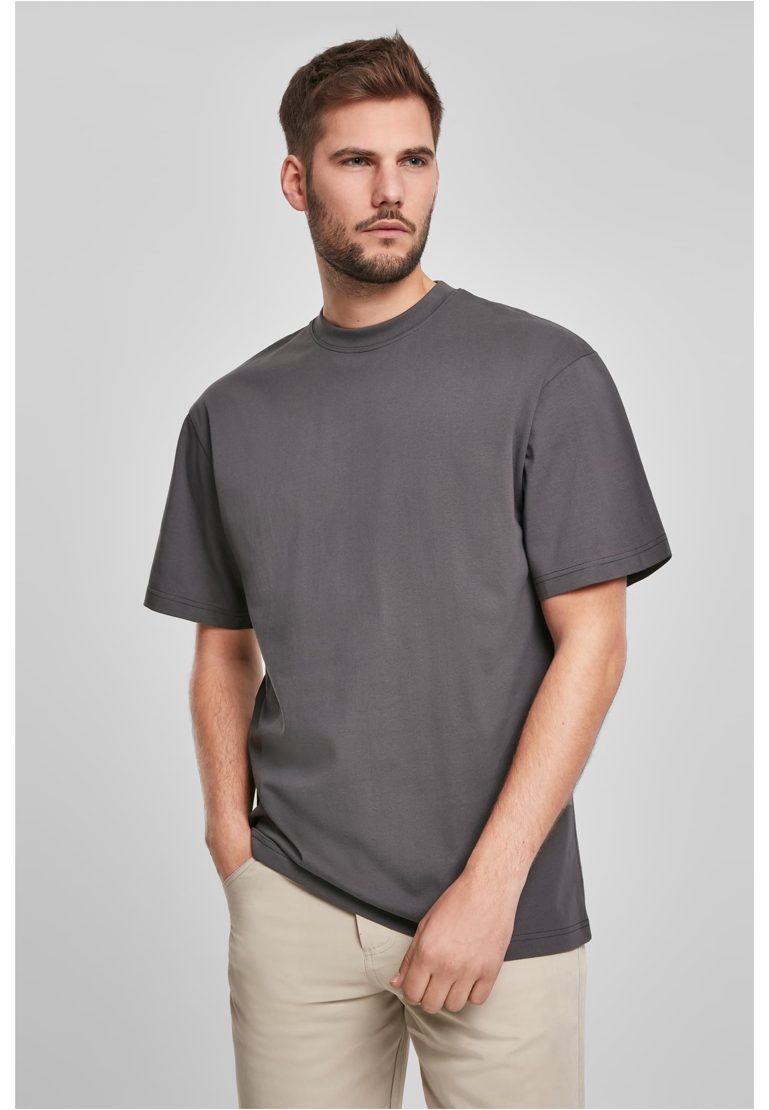 En man poserar i en mörkgrå Tall Tee med extra lång passform och beiga byxor mot en ljusgrå bakgrund.