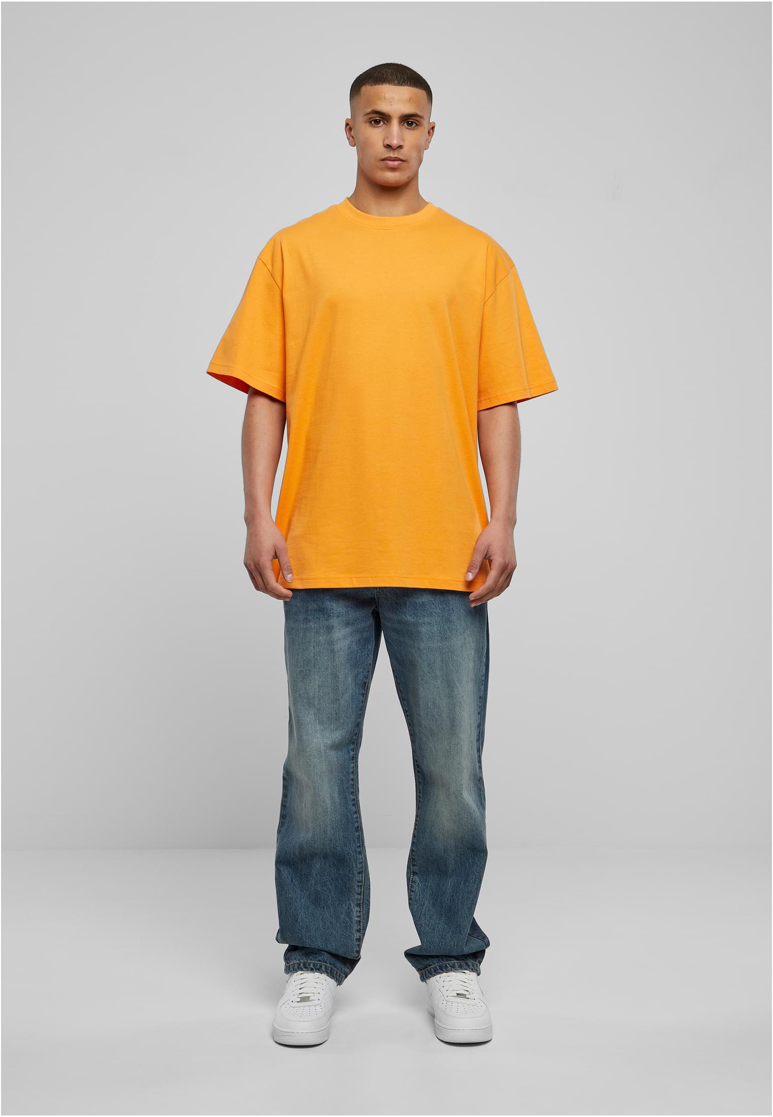 Frontbild på en manlig modell som bär en orange t-shirt, blå jeans och vita sneakers mot en ljusgrå bakgrund.