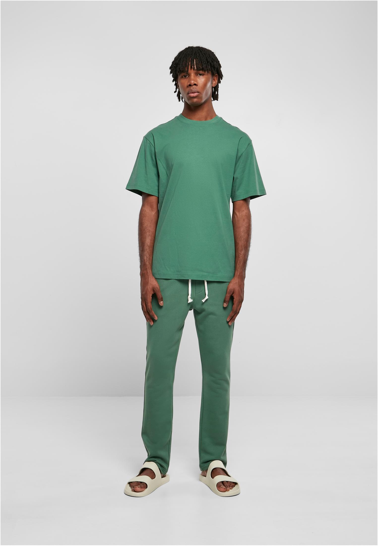 En man fotograferad framifrån klädd i en grön Tall Tee, matchande gröna mjukisbyxor och ljusa tofflor.