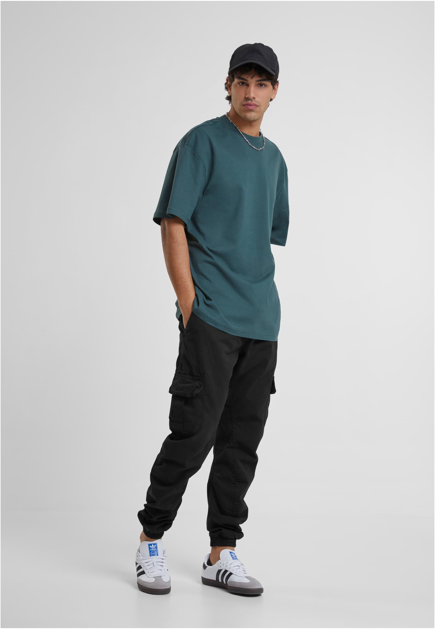 Manlig modell som bär en extra lång grönblå t-shirt, svarta cargobyxor och en svart keps mot en vit bakgrund.