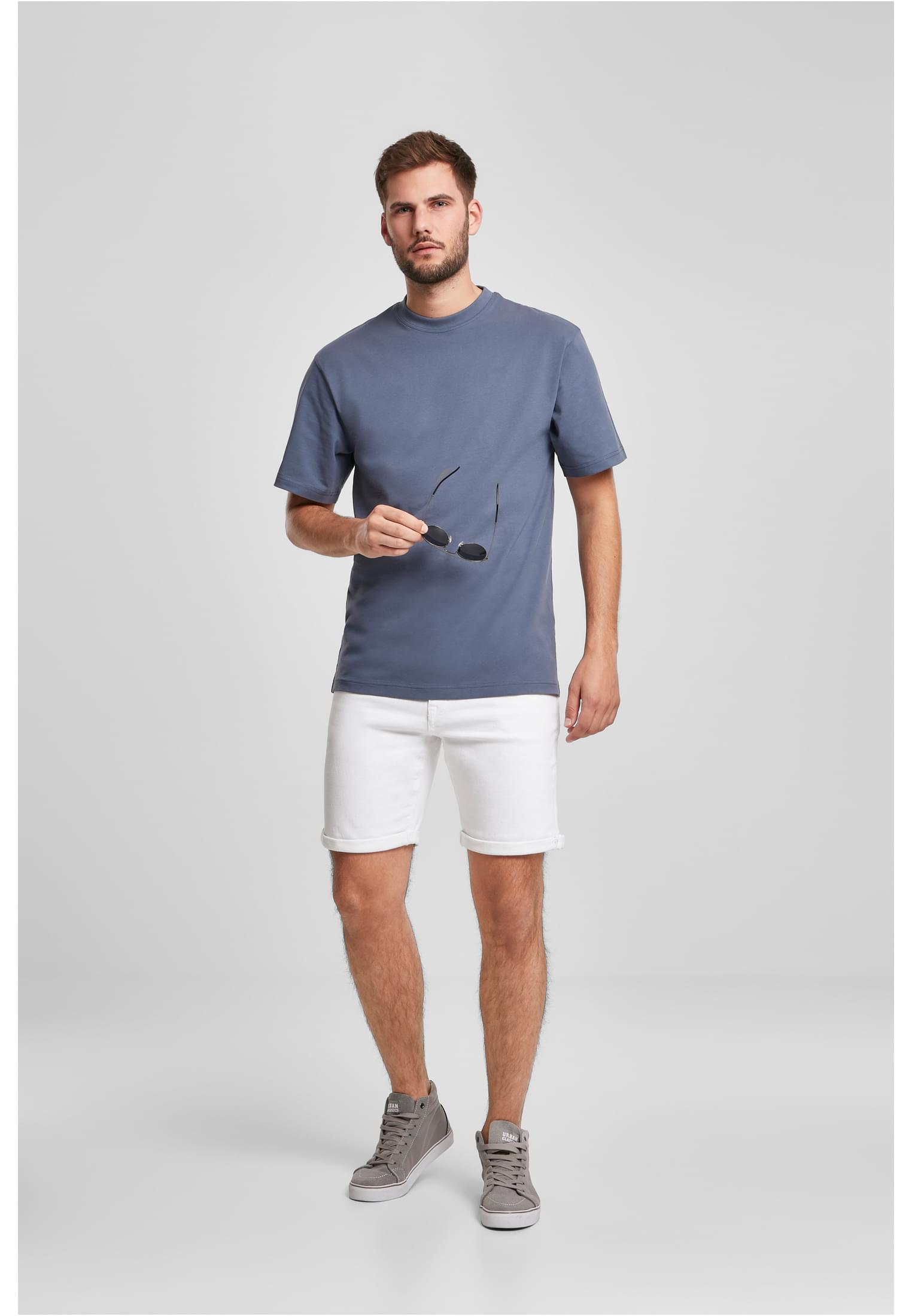 En manlig modell bär en mintgrön Tall Tee med extra lång passform, stylad med vita shorts och grå sneakers, samtidigt som han håller i ett par solglasögon.