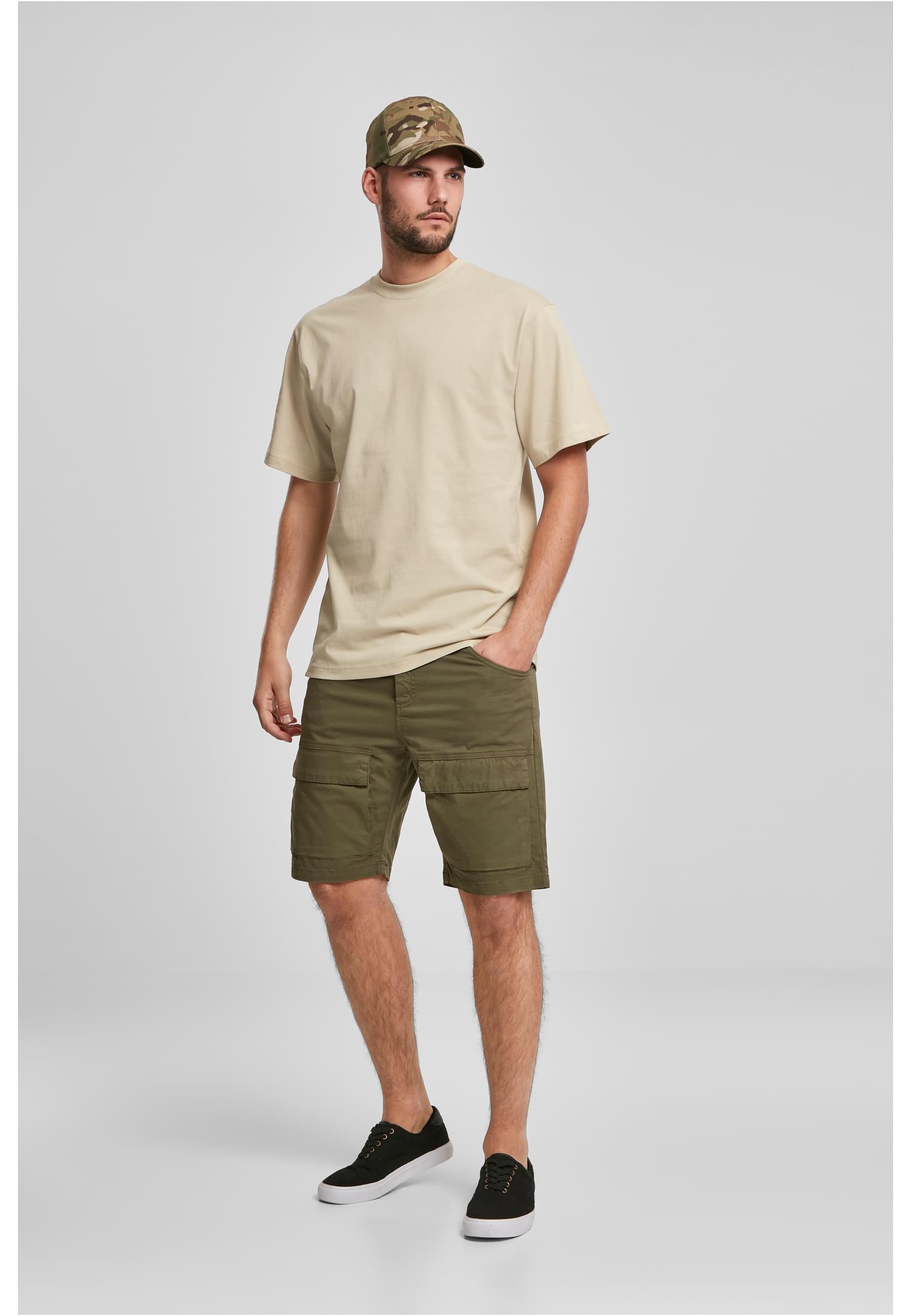 Manlig modell som bär en beige Tall Tee, olivgröna cargoshorts, en kamouflagekeps och svarta sneakers mot en ljusgrå studiobakgrund.