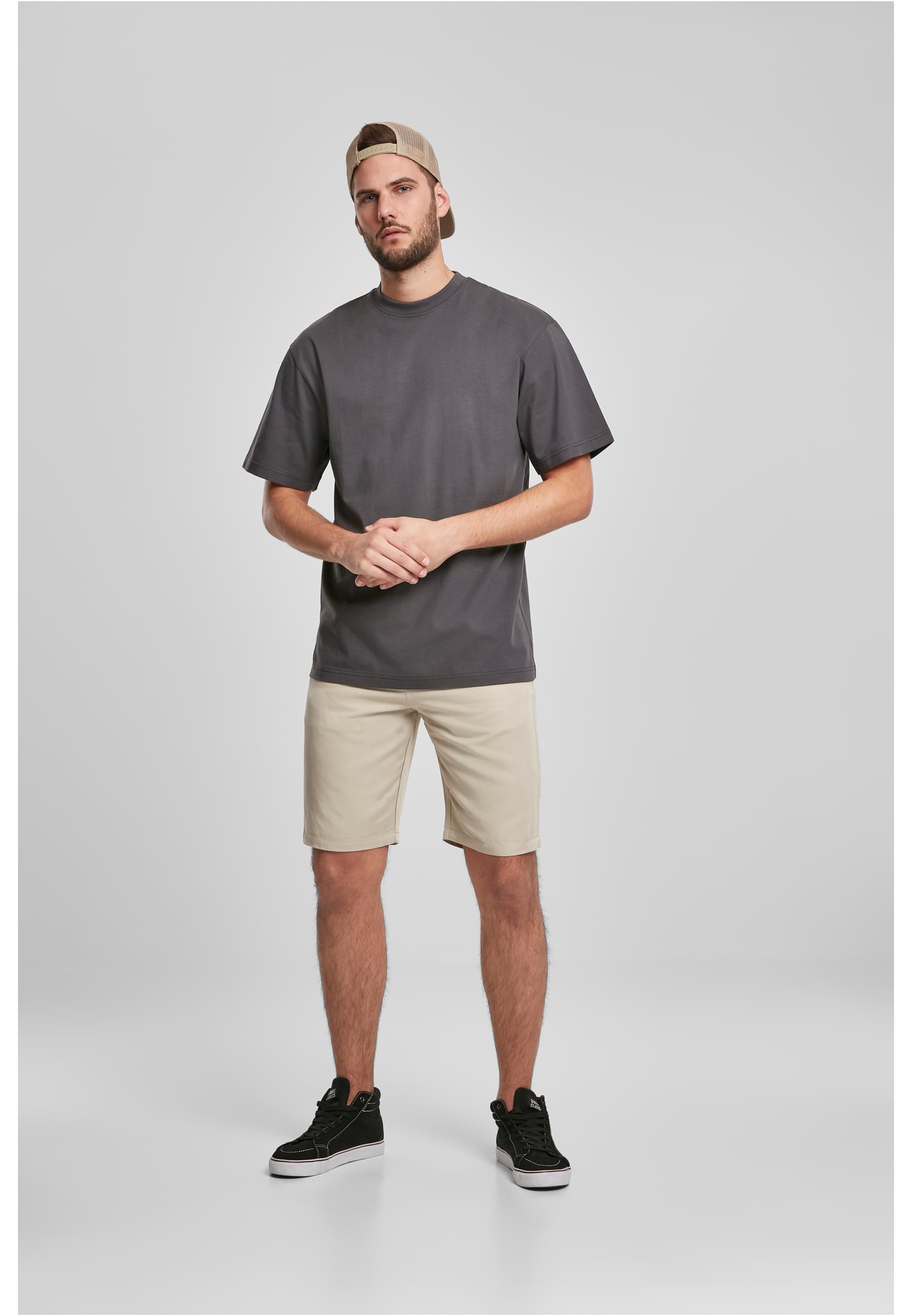 En man i helfigur som bär en mörkgrå Tall Tee, beiga shorts, svarta sneakers och en beige keps.