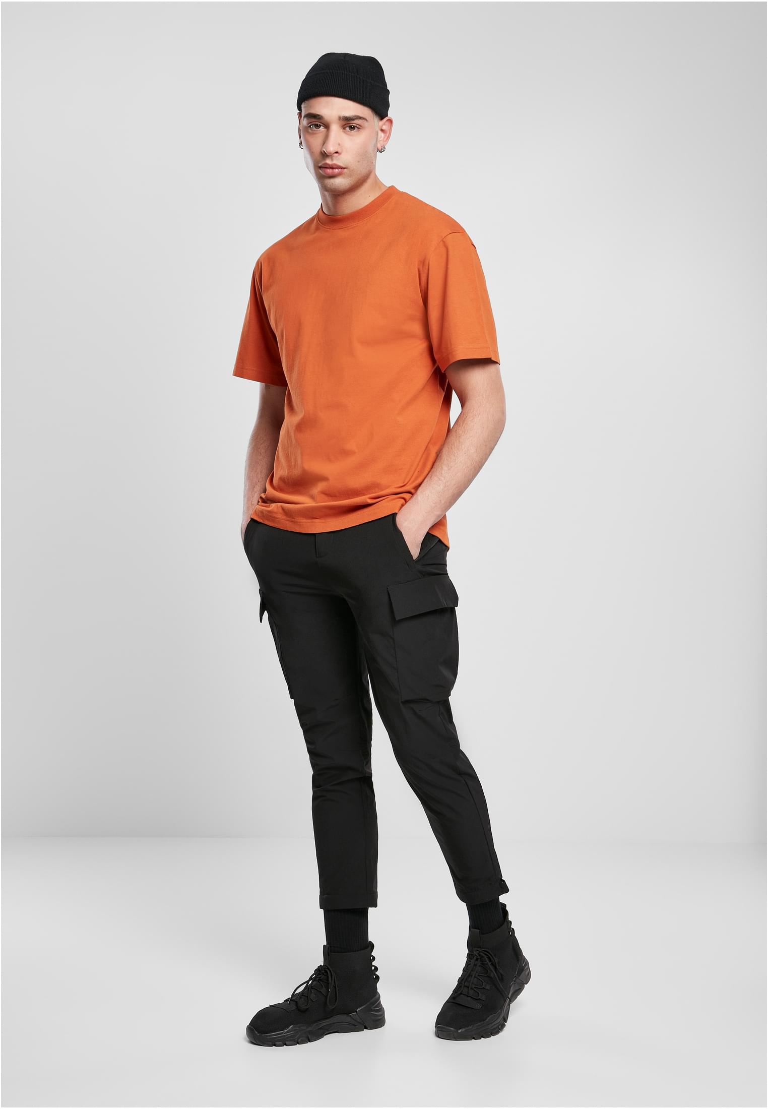 En manlig modell poserar i en orange Tall Tee, svarta cargobyxor och en svart mössa mot en ljusgrå bakgrund.