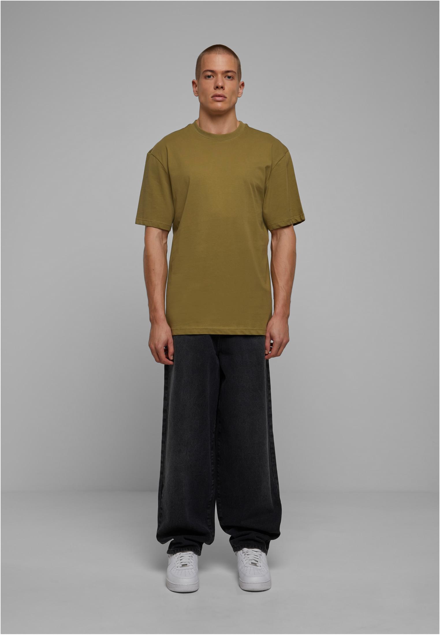 Manlig modell som bär en olivgrön UC Tall Tee med vida svarta jeans och vita sneakers, fotograferad framifrån mot en grå bakgrund.