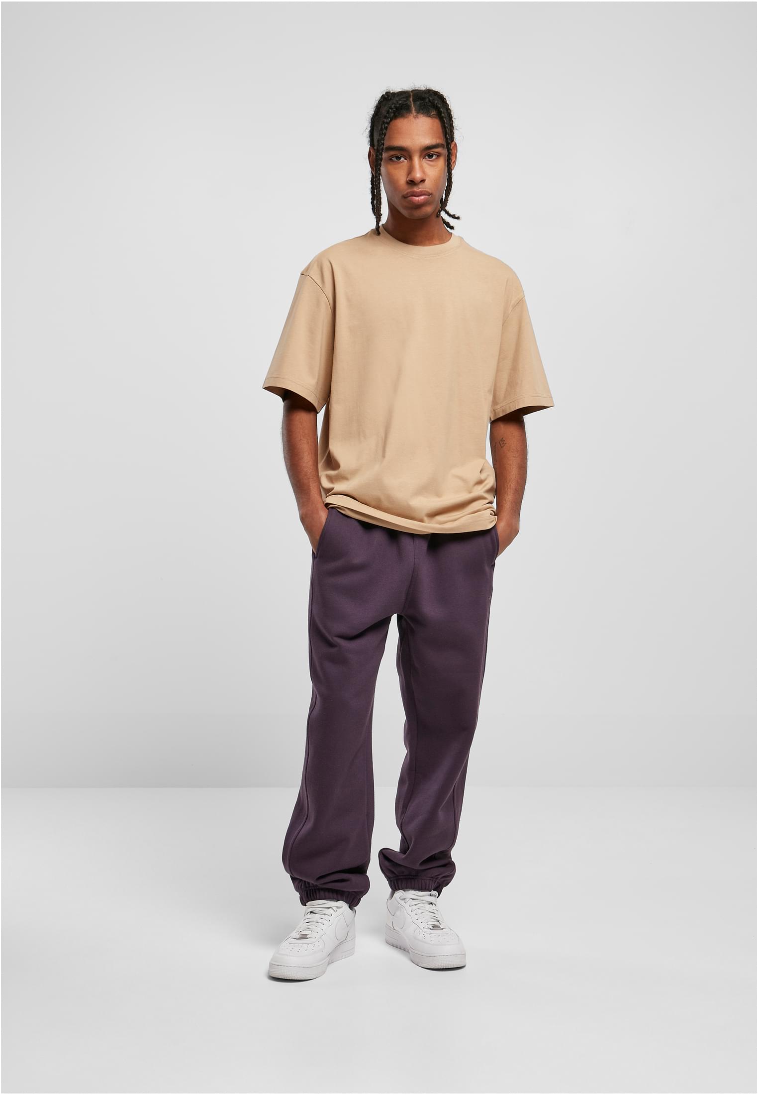 En manlig modell med flätat hår som bär en beige oversize t-shirt, mörklila mjukisbyxor och vita sneakers. Han står vänd mot kameran med händerna i fickorna mot en ljusgrå studiobakgrund.